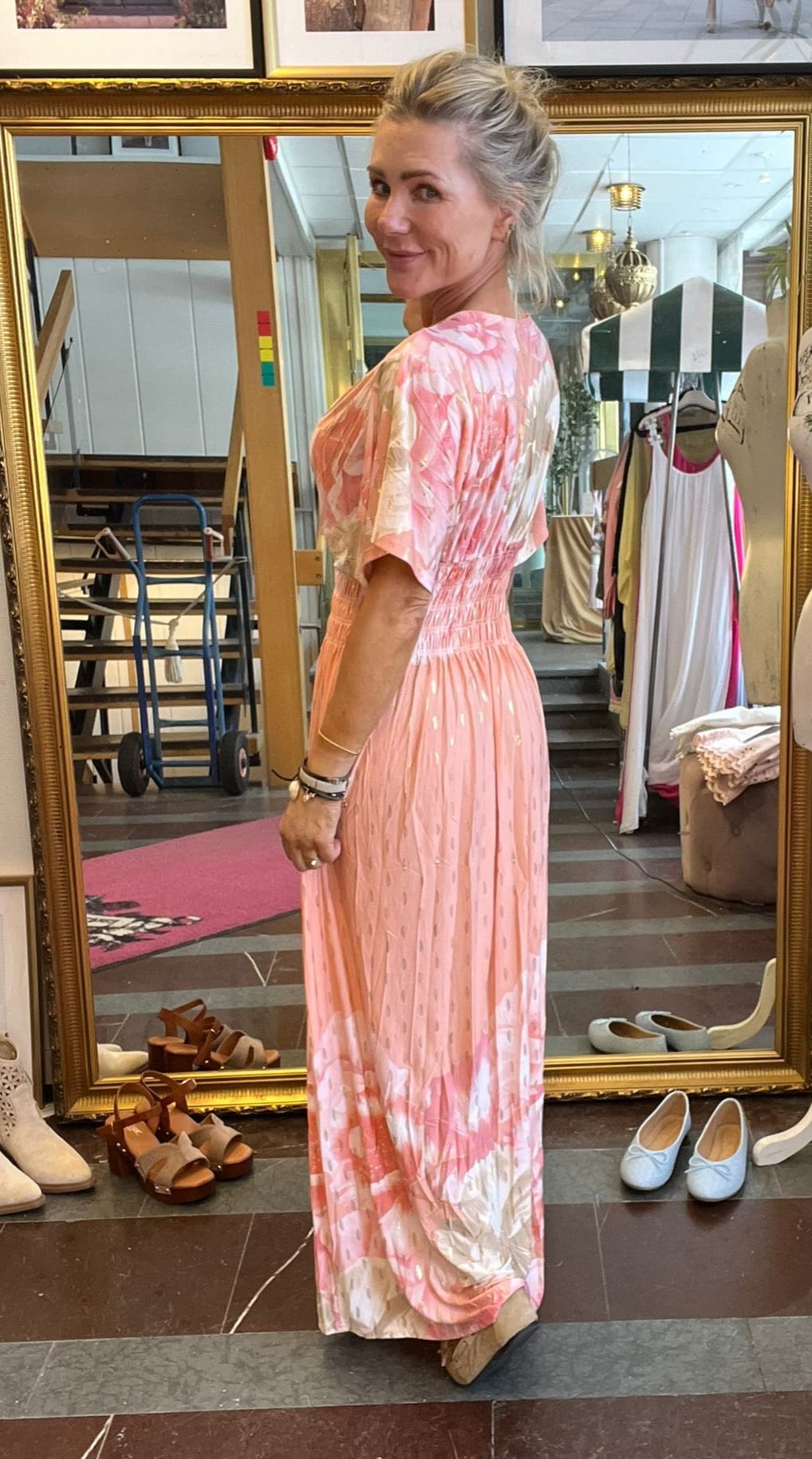 Blond kvinna står i en butik och visar upp en lång rosa blommig klänning, vänd i profil och ler mot kameran. Runt henne syns en stor guldramsspegel, skor på golvet och klädställningar.
