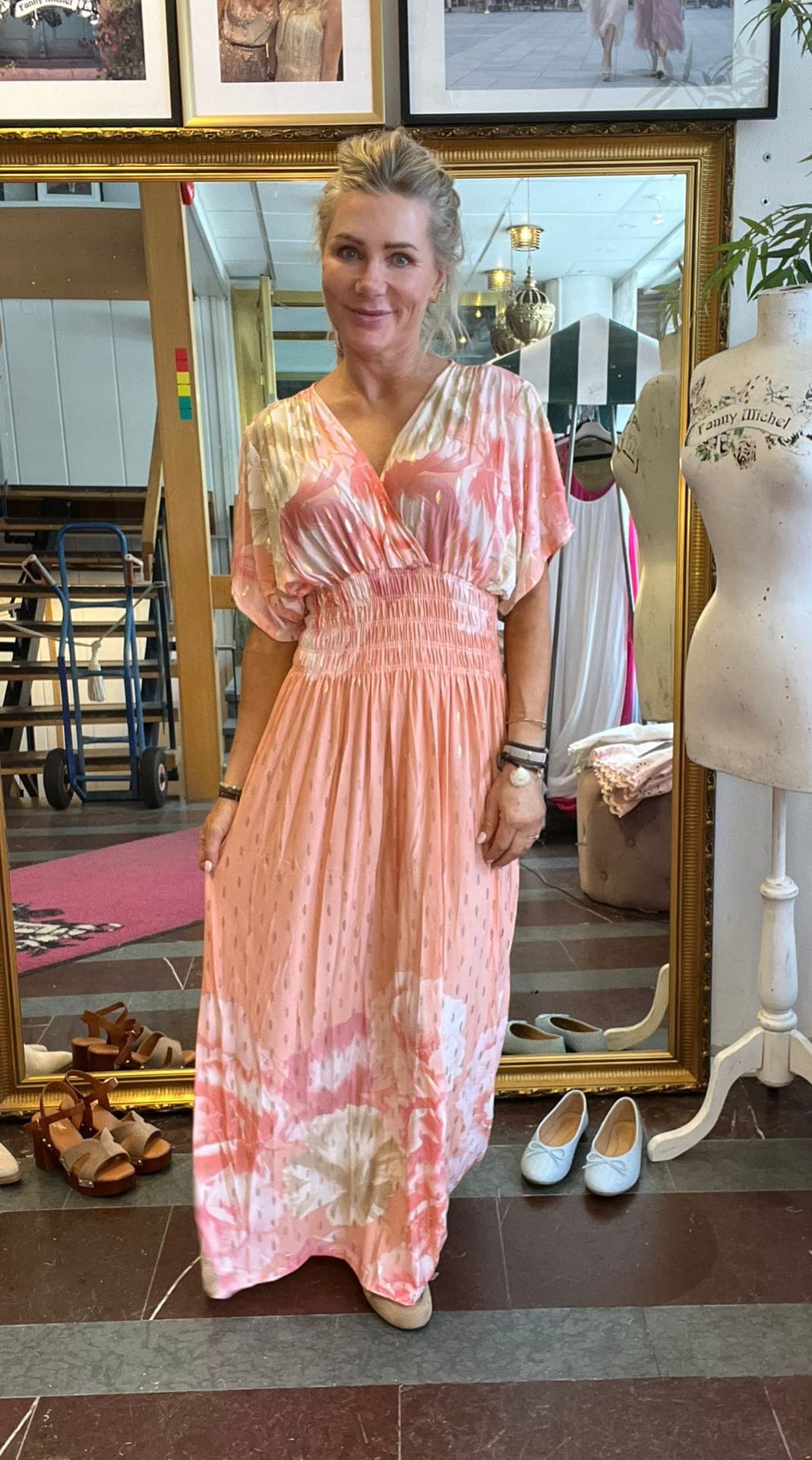 Kvinna står och poserar framför en stor guldramsspegel i en butik, klädd i lång rosa-och-vit mönstrad klänning med markerad midja. Runt henne syns skor på golvet och en provdocka med texten “Fanny Michel”.