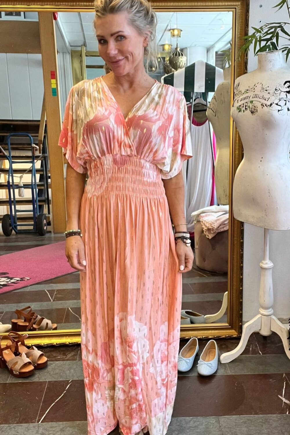 Blond kvinna står och ler i en klädbutik, iklädd en lång rosa- och vitmönstrad klänning med rynkad midja. Bakom henne syns en stor guldramsspegel, skor på golvet och en provdocka med texten “Fanny Michel”.