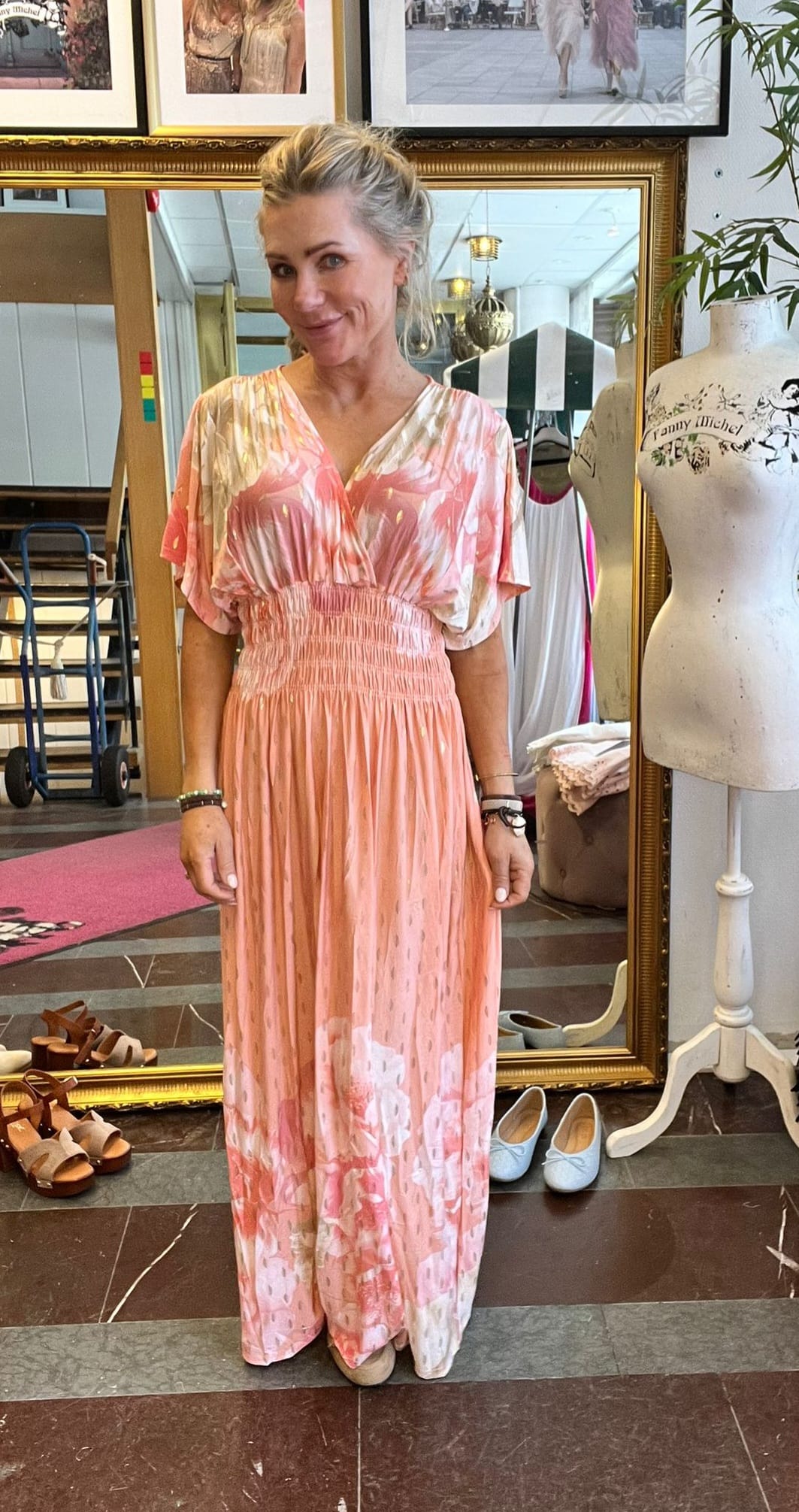 Blond kvinna står och ler i en klädbutik, iklädd en lång rosa- och vitmönstrad klänning med rynkad midja. Bakom henne syns en stor guldramsspegel, skor på golvet och en provdocka med texten “Fanny Michel”.