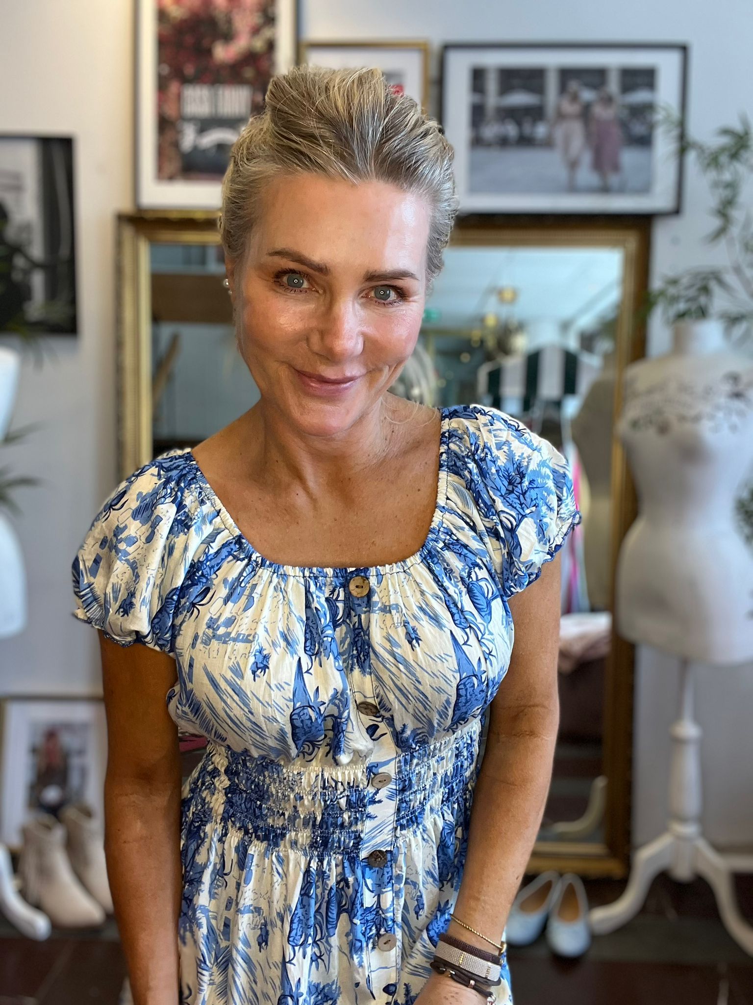 Leende kvinna med uppsatt blont hår lutar sig lätt fram mot kameran och bär en blåvit mönstrad klänning. I bakgrunden syns en inomhusmiljö med inramade foton, en spegel och växter.
