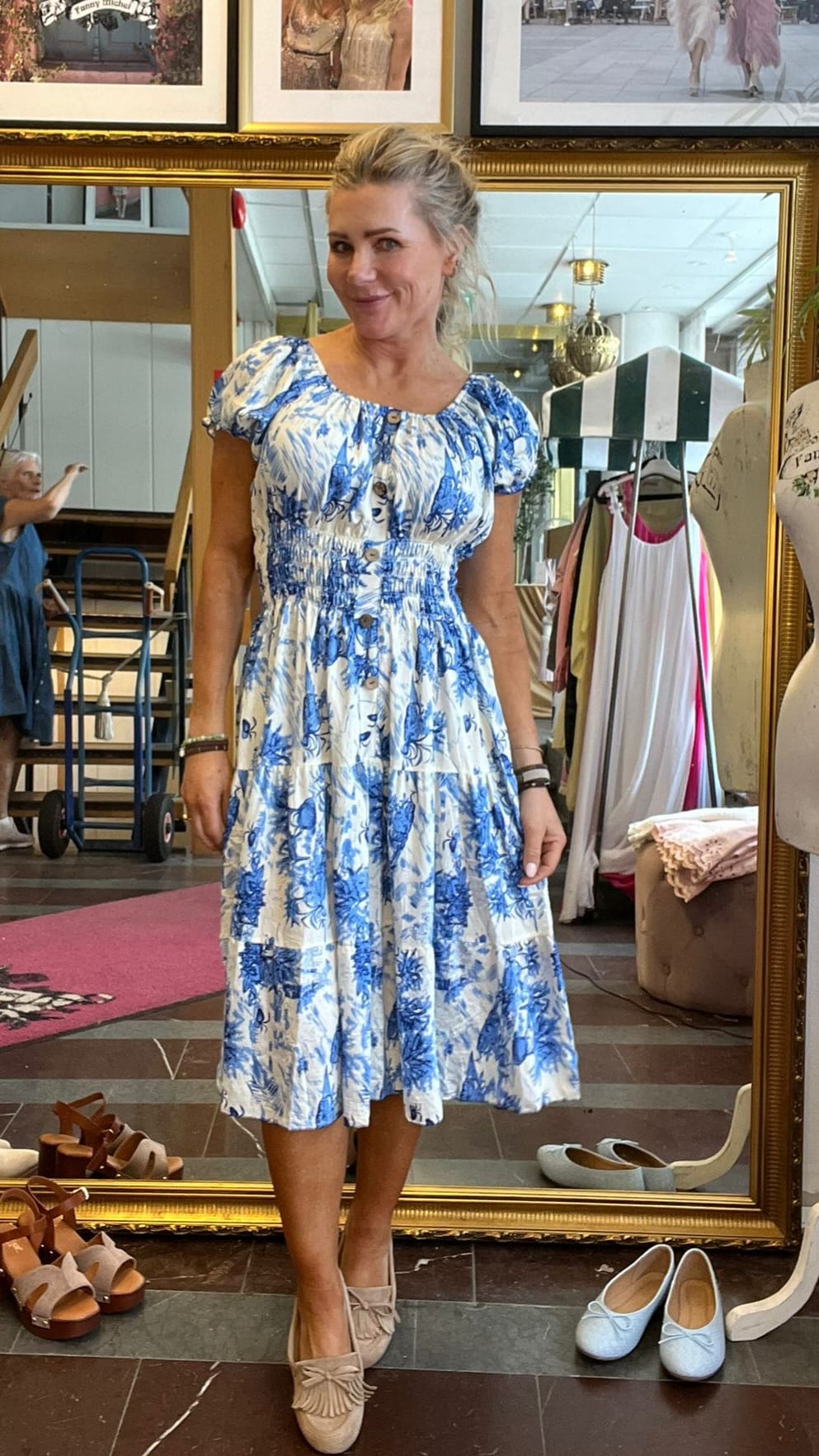 Kvinna står och poserar framför en stor guldramsspegel i en klädbutik, klädd i blåvit mönstrad sommarklänning och beige skor. Klädställningar, skyltdocka och flera par skor syns runt henne.