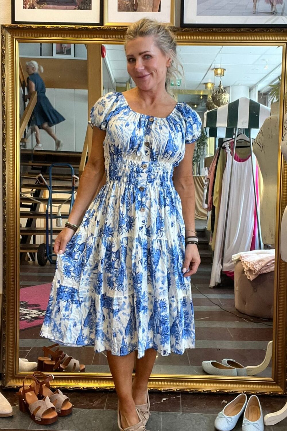 Blond kvinna står framför en stor guldramsspegel och håller ut kjolen på en vit klänning med blått blommönster. Hon provar outfit i en butik, med skor på golvet och kläder på ställning bakom.