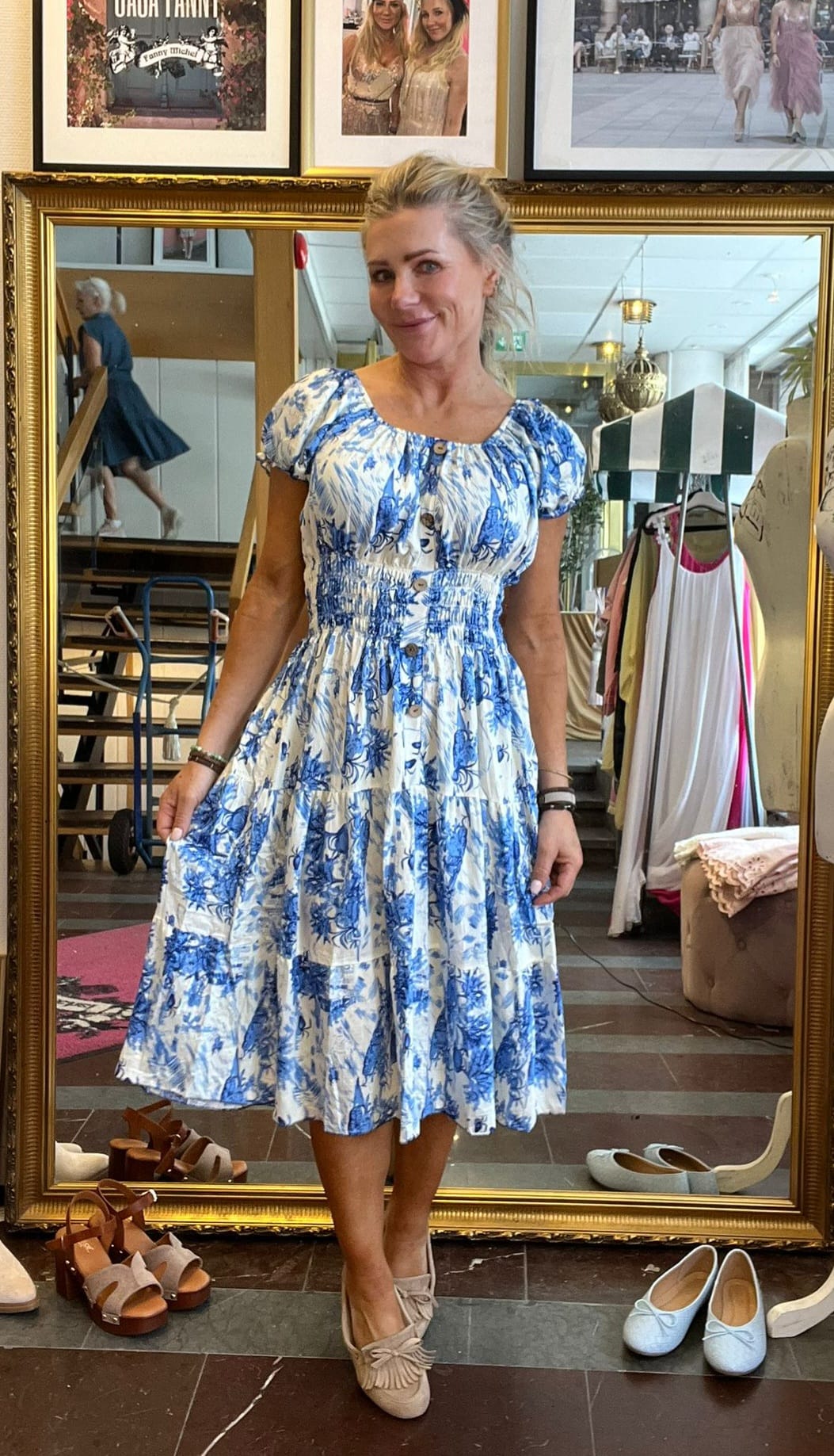 Blond kvinna står framför en stor guldramsspegel och håller ut kjolen på en vit klänning med blått blommönster. Hon provar outfit i en butik, med skor på golvet och kläder på ställning bakom.