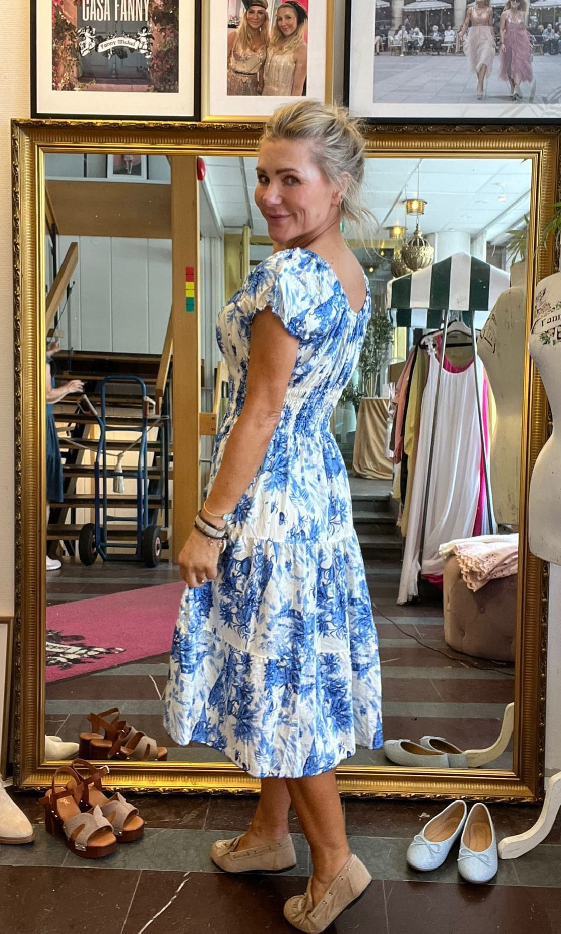 Kvinna står och ler mot kameran medan hon provar en vit klänning med blått blommönster, fotograferad från sidan. Hon är i en butik med stor guldramad spegel, skor på golvet och affischer ovanför. Text: ”CASA FANNY”.