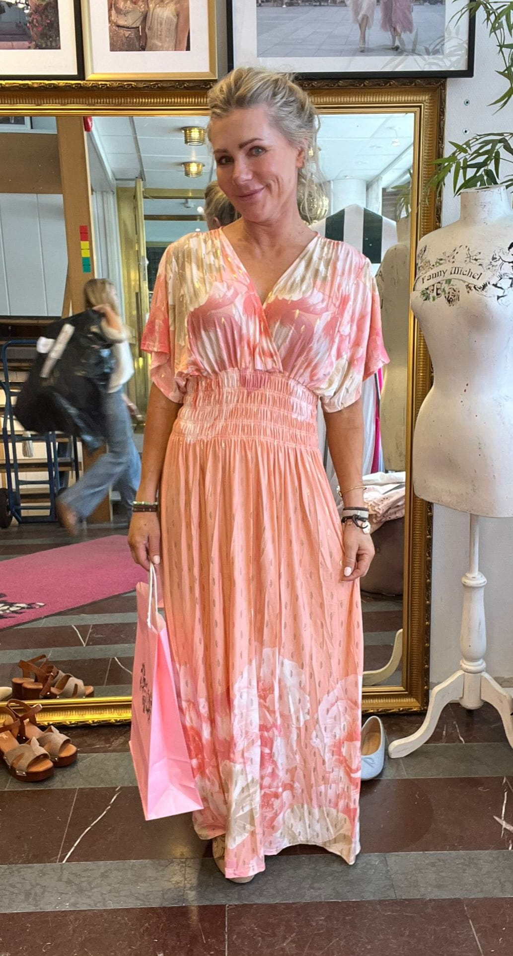 Leende kvinna står i en klädbutik och håller en rosa shoppingpåse, iklädd lång rosa- och vitmönstrad klänning med markerad midja. Bakom henne syns en stor spegel, skor på golvet och en provdocka med texten “Tommy Michel”.