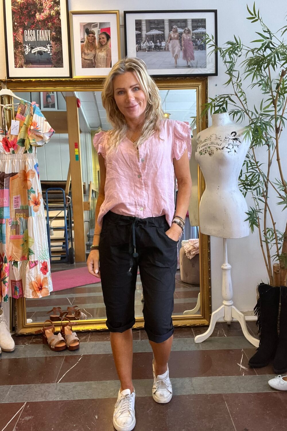 Leende kvinna står framför en stor guldramsspegel i en butik, med händerna i fickorna. Runt henne syns en färgglad klänning på galge, skor, en provdocka och inramade foton; text: “CASA FANNY”.