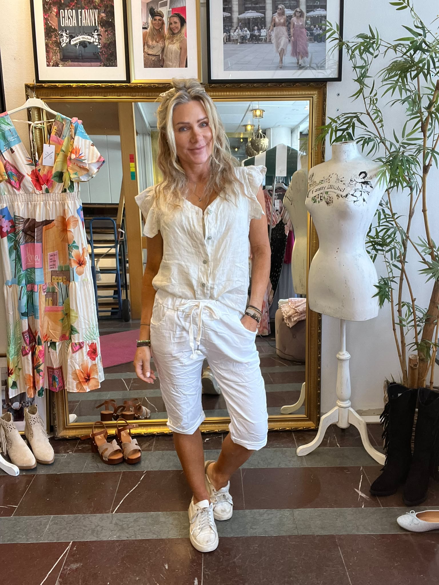 Kvinna i vit kortärmad blus och vita capribyxor står och ler i en klädbutik, framför en stor guldramad spegel. Intill syns en färgstark klänning och en provdocka med texten “Fanny Michel”.