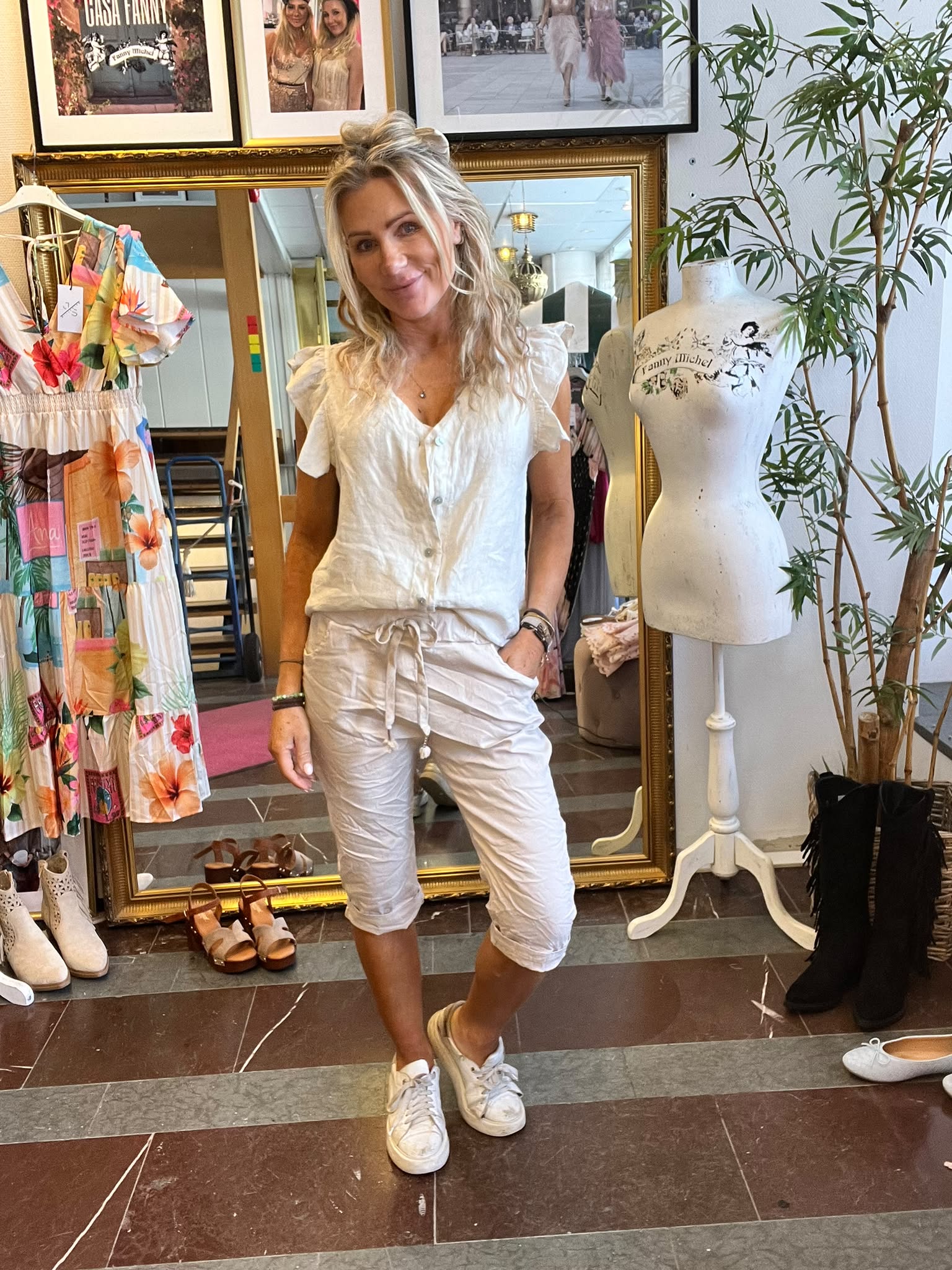 Blond kvinna i vit blus och ljusa knälånga byxor poserar med handen i fickan i en klädbutik. Bredvid står en provdocka med texten ”Fanny Michel”, bakom syns stor guldramsspegel, klänning och skor.