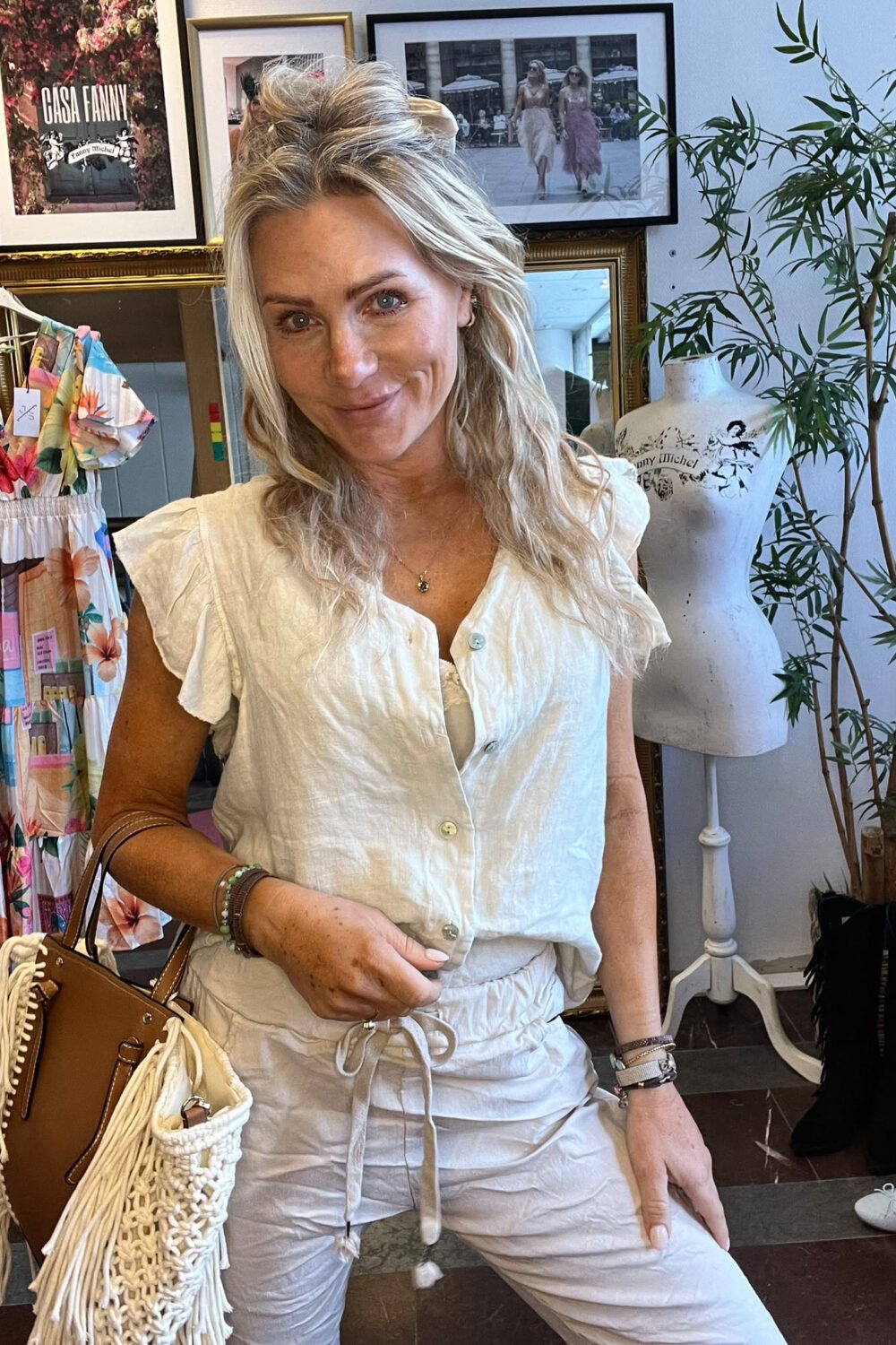 Blond kvinna i vit blus och ljusa byxor ler mot kameran och håller en fransad väska. Hon står i en butik/ateljé med färgglad klänning, provdocka och inramade bilder; text på tavla: “CASA FANNY”.
