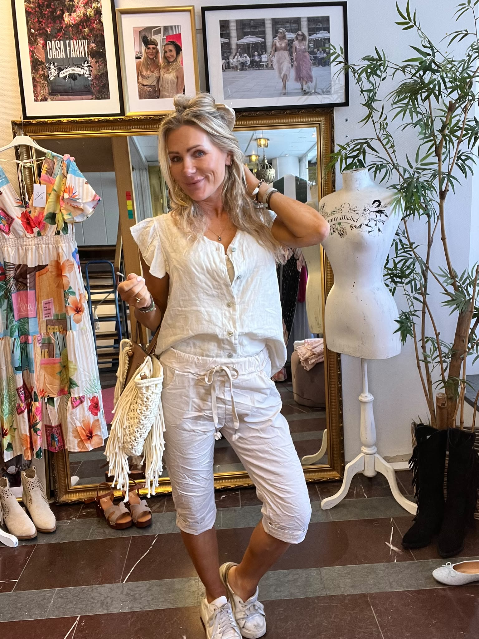 Leende kvinna i vit kortärmad blus och ljusa byxor poserar med handen i håret i en klädbutik. Runt henne syns färgglad klänning, skor och skyltdocka; på väggen hänger foton med texten “CASA FANNY”.