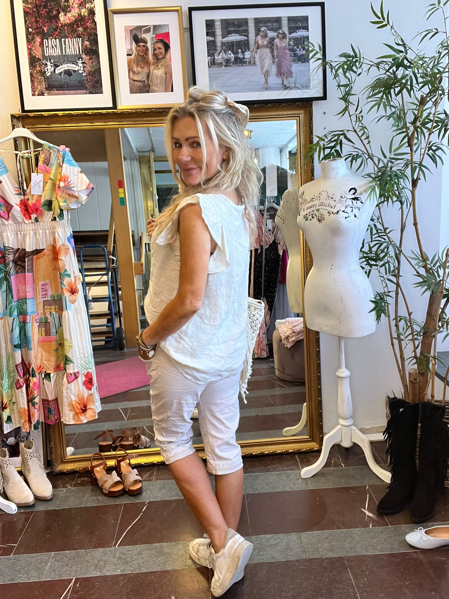 Blond kvinna i vit topp och vita capribyxor poserar och ler mot kameran i en klädbutik. Bakom henne står en stor guldspegel, en provdocka med texten “Fanny Michel”, färgglad klänning och inramade foton.