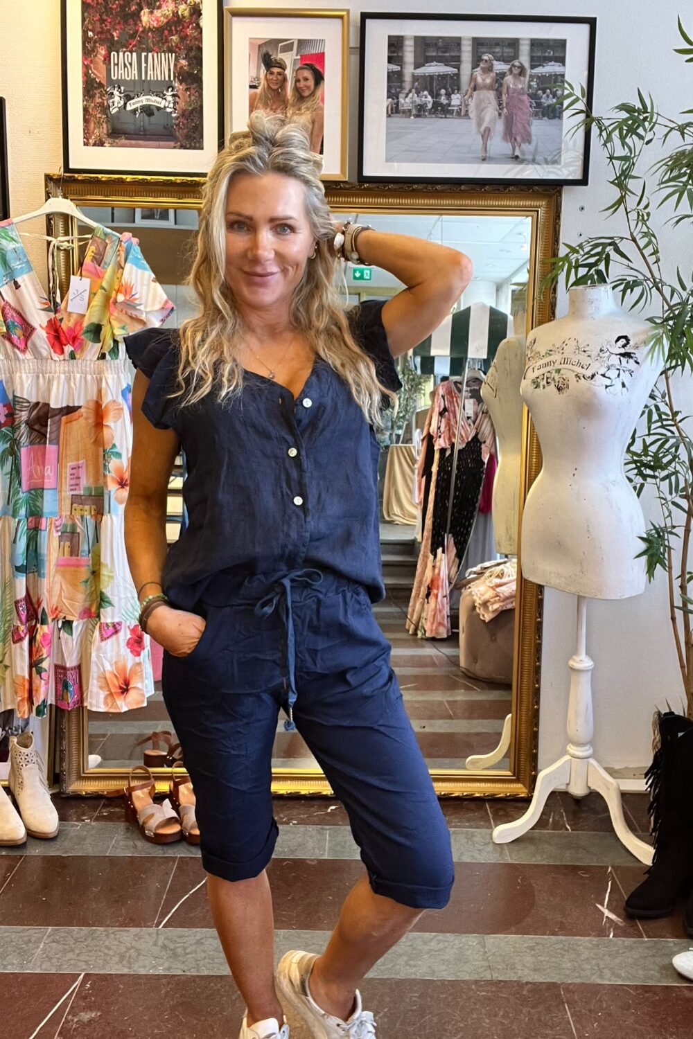 Leende kvinna i mörkblå jumpsuit poserar med ena handen i fickan och den andra bakom huvudet i en klädbutik, framför en guldramsspegel, klädställningar och provdocka. Inramad text på väggen: “CASA FANNY”.