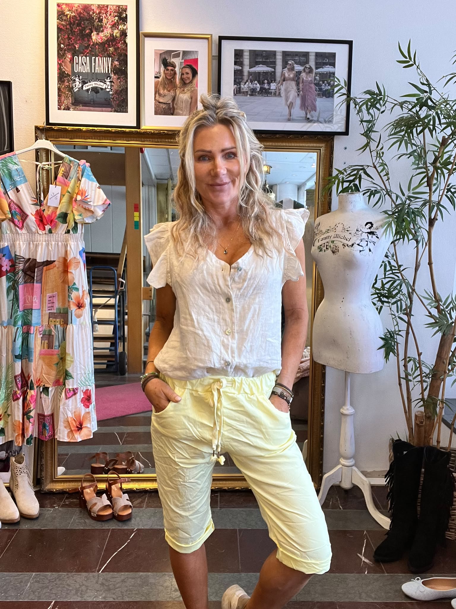 Leende kvinna står med händerna i fickorna i en klädbutik, klädd i vit blus och ljusgula shorts. Bakom syns spegel, klänning på galge, provdocka och inramade foton med texten ”CASA FANNY”.