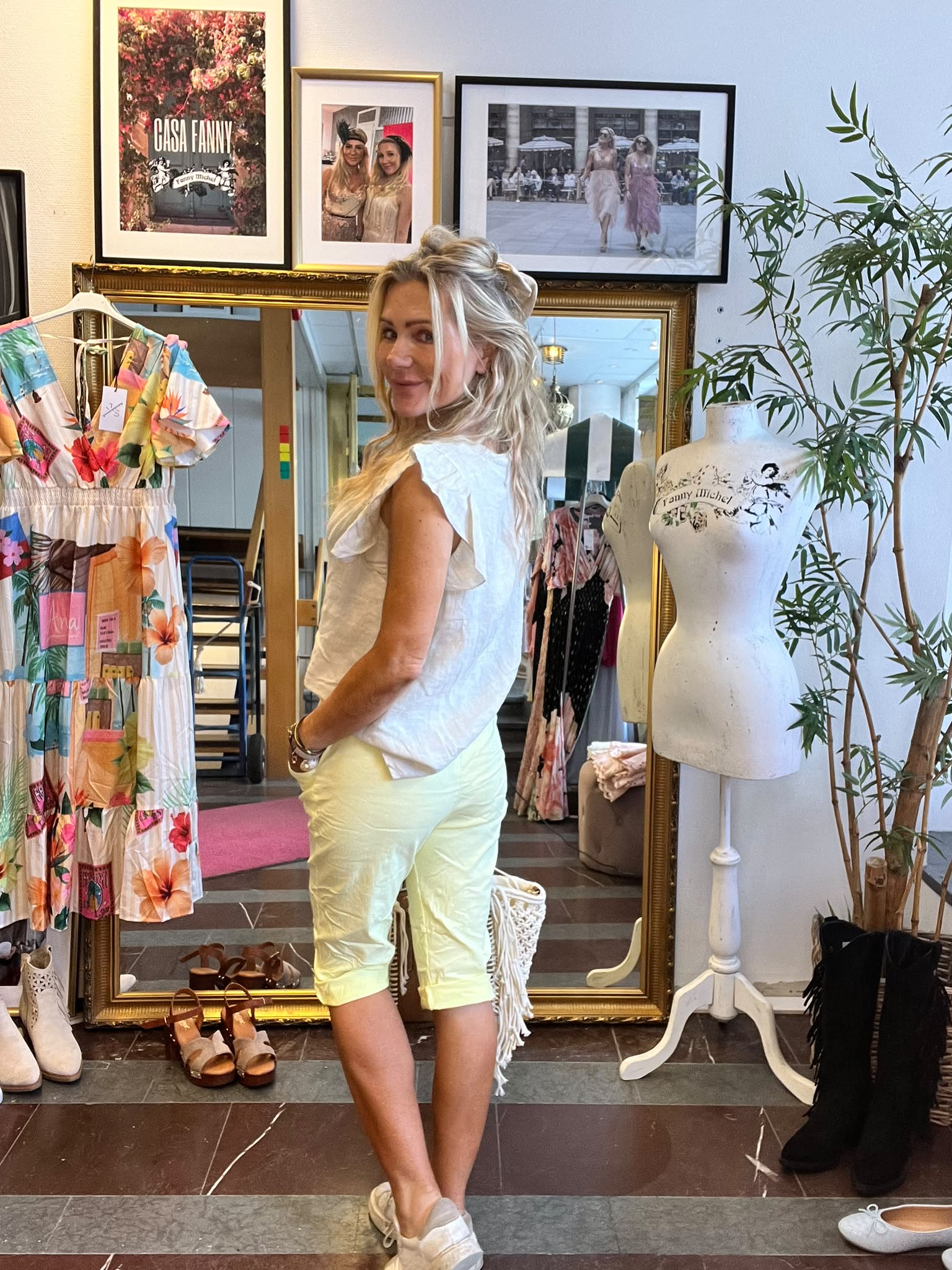 Leende kvinna poserar framför en stor guldramsspegel i en klädbutik, klädd i vit topp och ljusa shorts. Runt henne syns färgglad klänning på galge, skyltdocka och väggbilder med texten “CASA FANNY”.