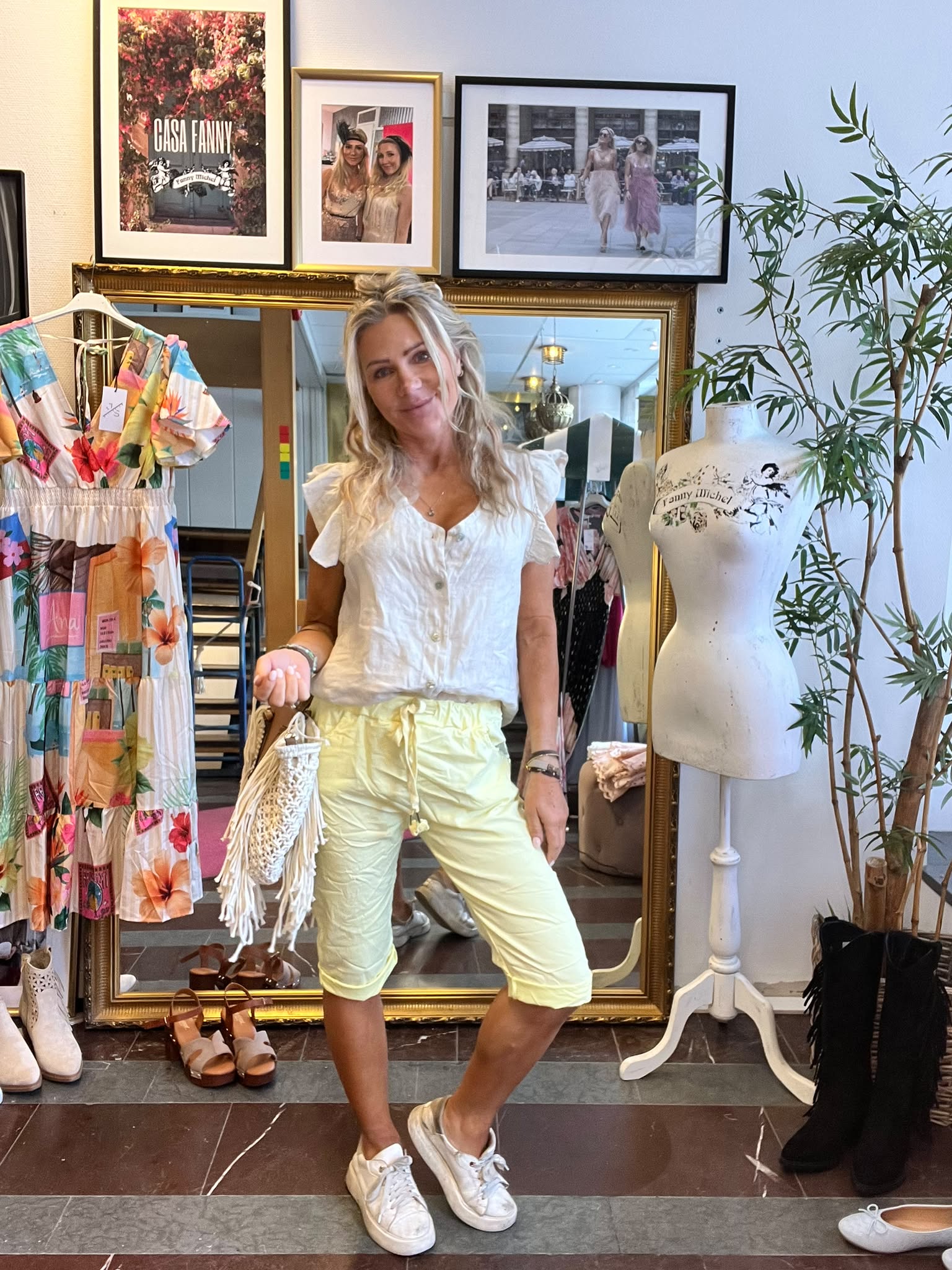 Kvinna poserar och ler i en klädbutik, klädd i vit blus och ljusgula shorts och håller en fransväska. Bredvid står en provdocka och en färgglad klänning; på väggen syns texten ”CASA FANNY”.