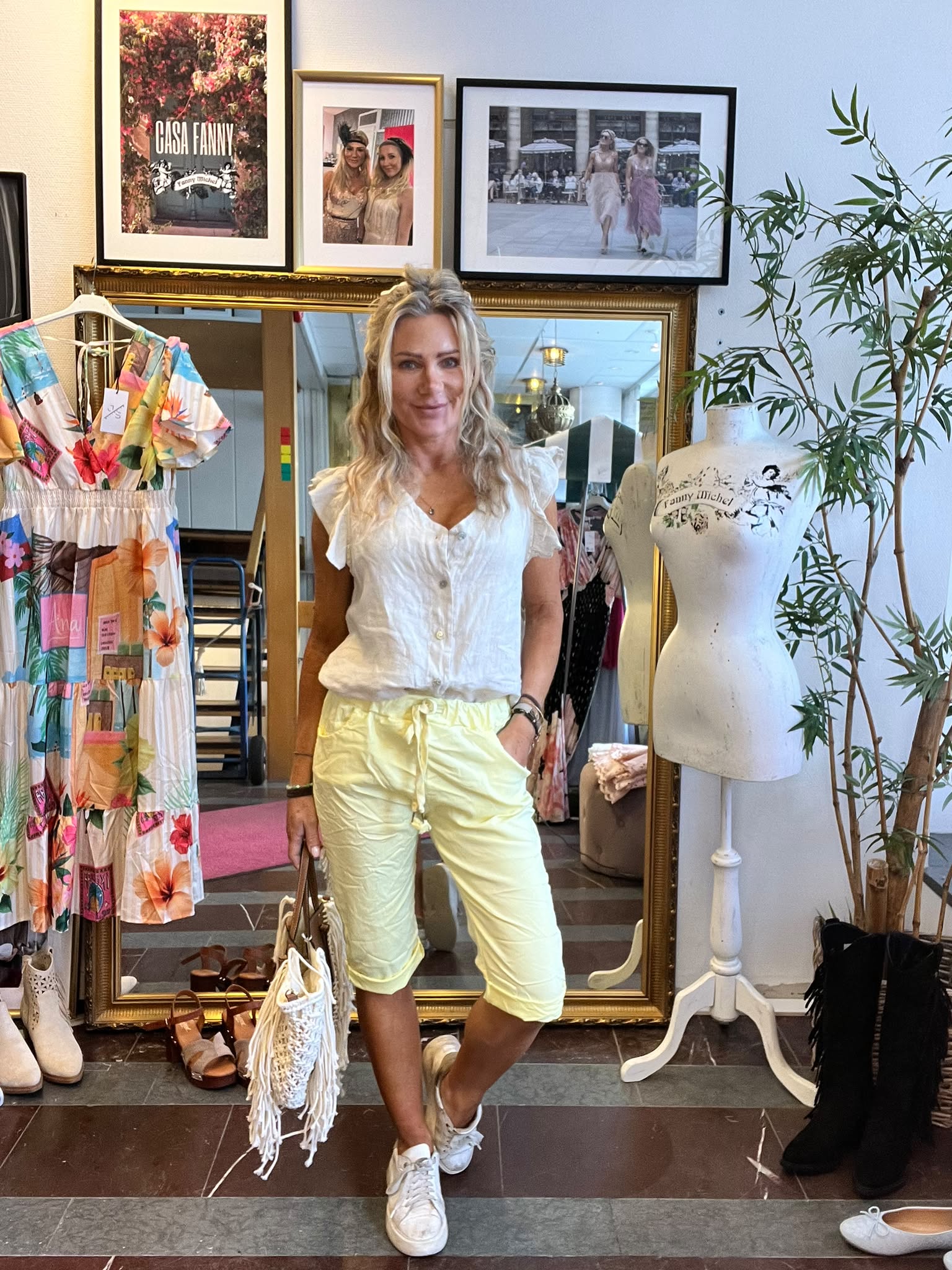 Blond kvinna poserar leende i en klädbutik framför en stor guldramsspegel, klädd i vit blus och ljusgula shorts, med handväska. Intill står en provdocka med texten “Fanny Michel” och en färgglad klänning; tavla visar “CASA FANNY”.