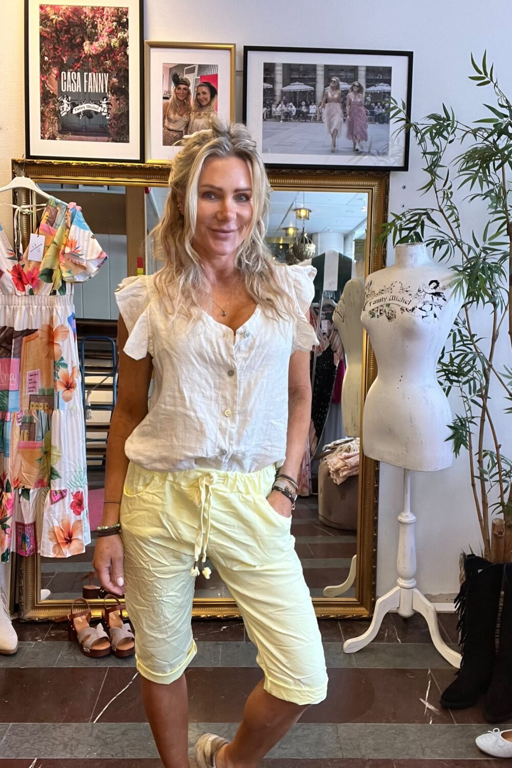 Leende kvinna står framför en stor guldramsspegel i en klädbutik, med handen i fickan. Bredvid syns en färgglad klänning och en provdocka. På väggen hänger tavlor med texten “CASA FANNY”.