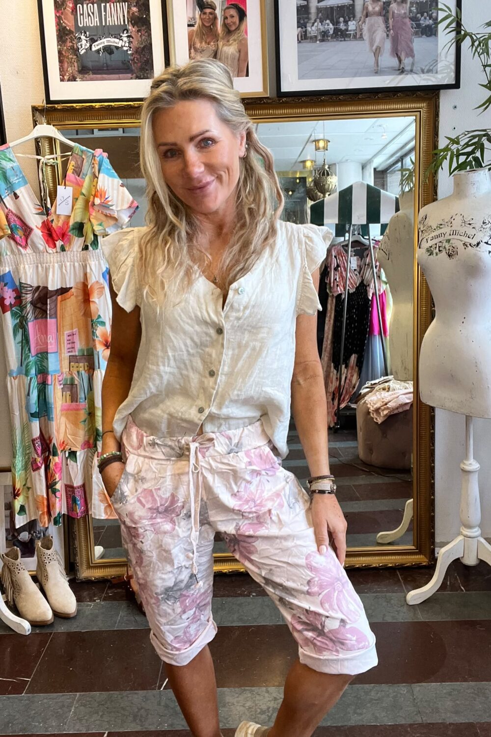 Leende blond kvinna poserar med händerna i fickorna framför en stor spegel i en klädbutik, omgiven av färgglad klänning och provdocka. På väggen syns texten “CASA FANNY” och “Fanny Michel”.