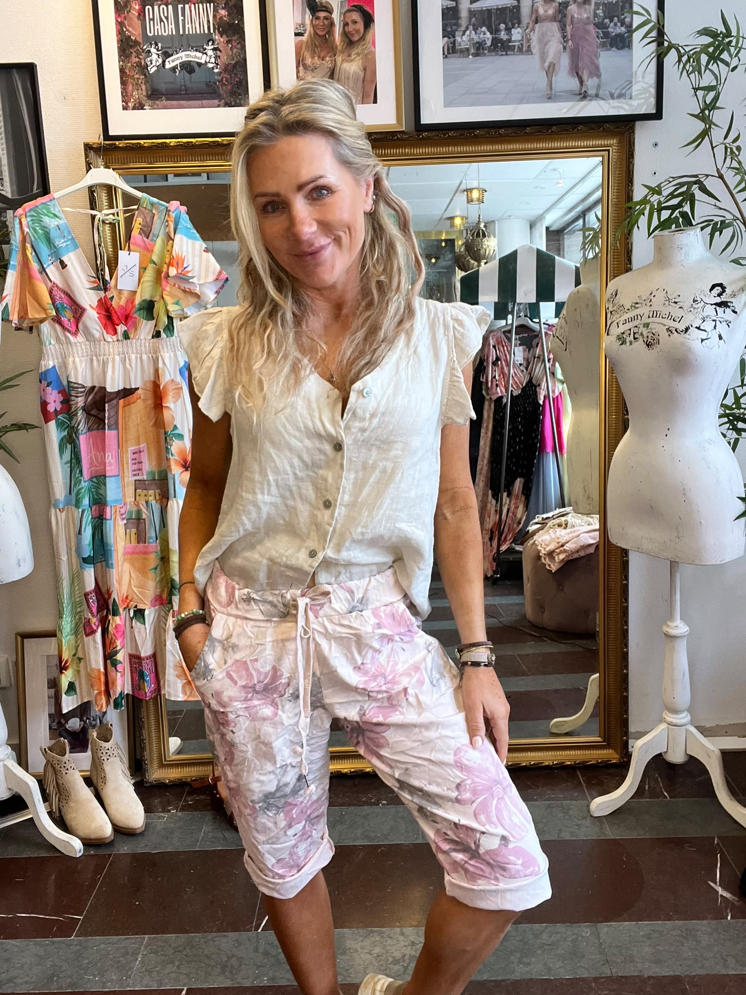 Leende blond kvinna poserar med händerna i fickorna framför en stor spegel i en klädbutik, omgiven av färgglad klänning och provdocka. På väggen syns texten “CASA FANNY” och “Fanny Michel”.