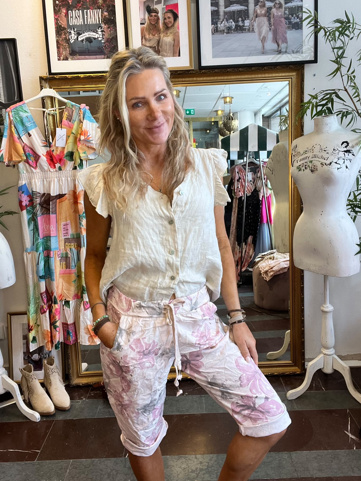 Leende kvinna står med handen i fickan och poserar i en klädbutik, klädd i vit blus och rosa blommiga shorts. Bakom syns klänningar, skyltdocka med texten “Fanny Michel” och tavla med “CASA FANNY”.