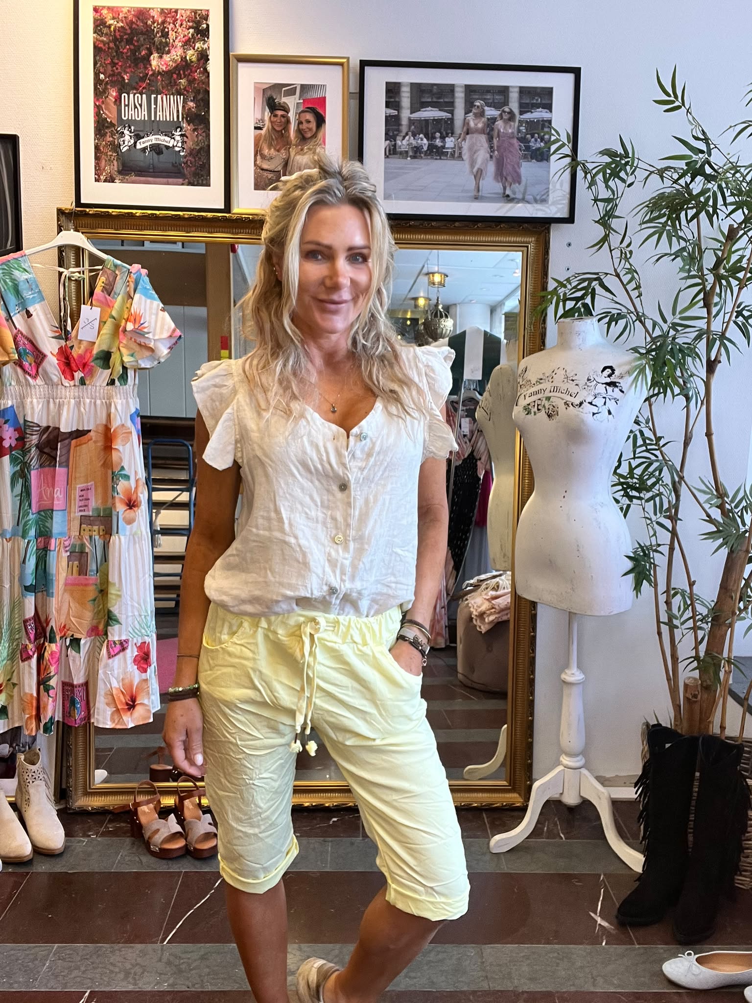 Leende kvinna står framför en stor guldramsspegel i en klädbutik, med handen i fickan. Bredvid syns en färgglad klänning och en provdocka. På väggen hänger tavlor med texten “CASA FANNY”.
