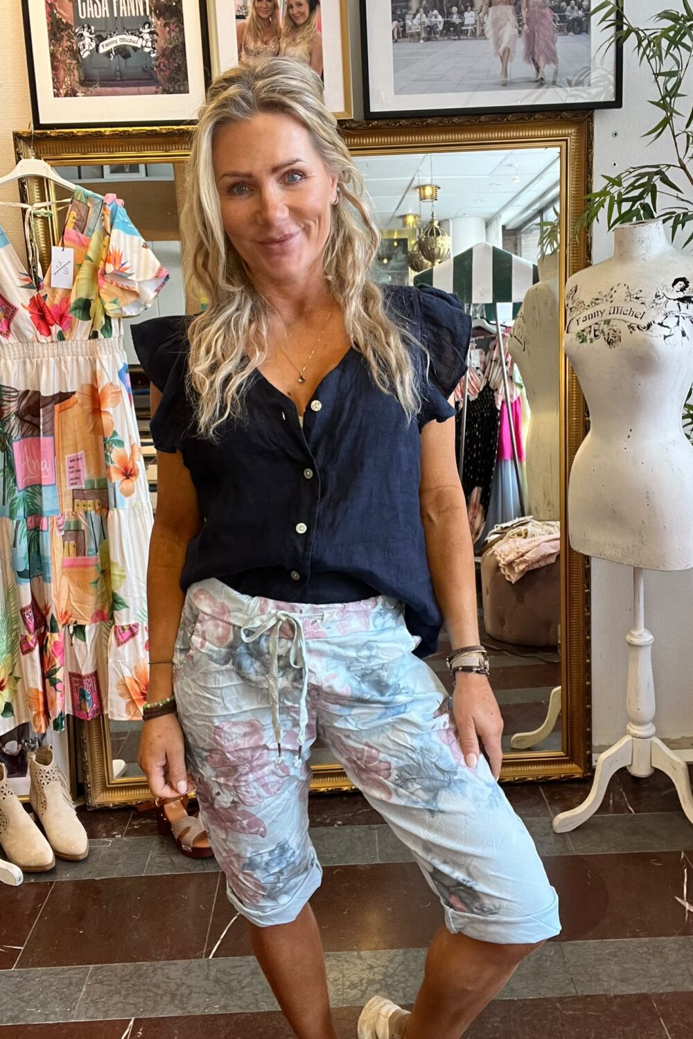 Kvinna med långt blont hår poserar och ler i en klädbutik, klädd i mörk blus och ljusa mönstrade capribyxor. Bredvid syns en färgglad klänning och provdocka med texten “Fanny Michel”.