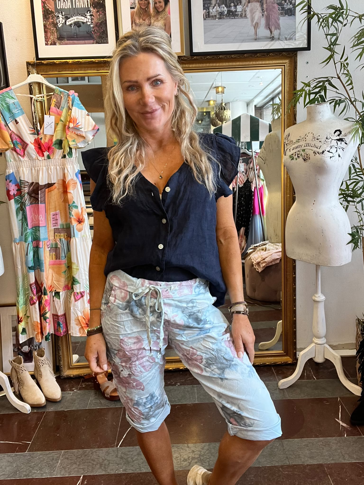 Kvinna med långt blont hår poserar och ler i en klädbutik, klädd i mörk blus och ljusa mönstrade capribyxor. Bredvid syns en färgglad klänning och provdocka med texten “Fanny Michel”.