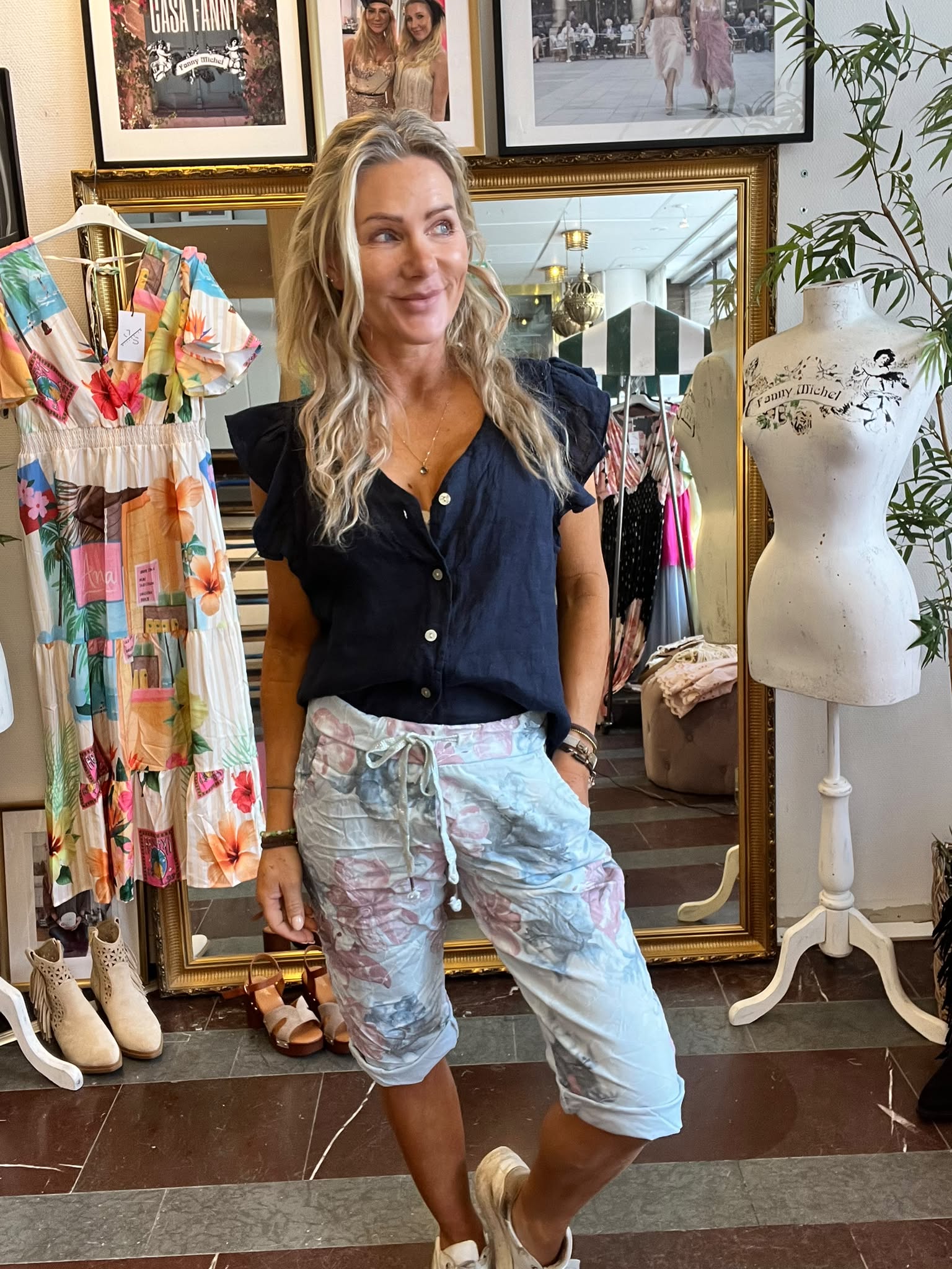 Leende kvinna står avslappnat och tittar åt sidan i en klädbutik, framför en stor guldramsspegel. Bredvid syns en färgglad klänning på galge och en provdocka med texten “Fanny Michel”.