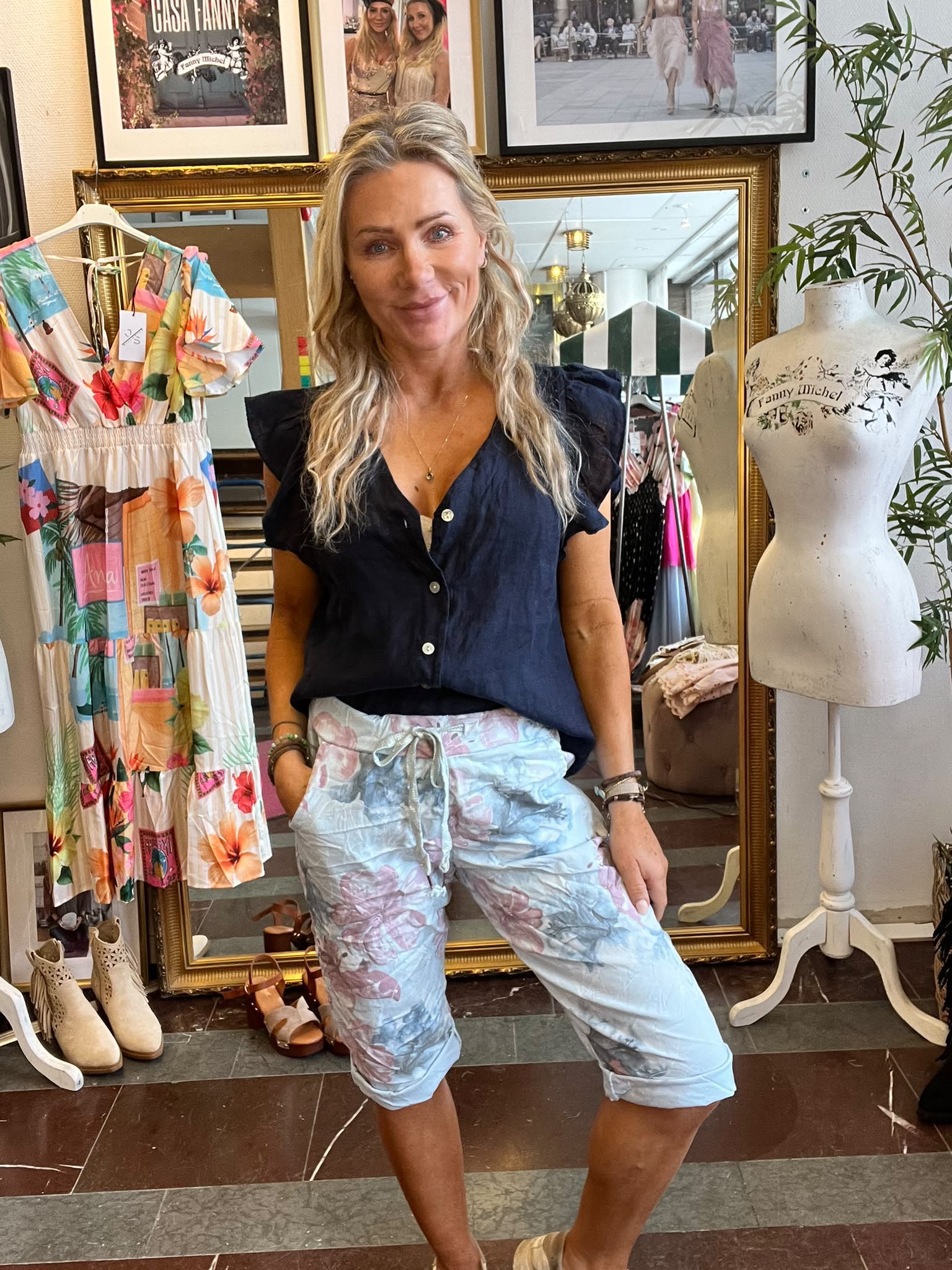 Blond kvinna står och ler i en klädbutik, iklädd mörkblå blus och ljusa mönstrade byxor. Hon poserar framför en guldramsspegel, med en färgglad klänning på galge och en provdocka märkt “Fanny Michel”.