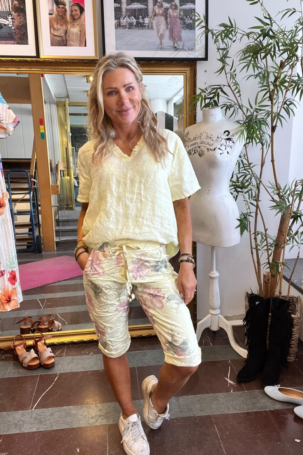 Blond kvinna står avslappnat och ler i en klädbutik, med händerna i fickorna, ljusgul topp och blommiga shorts. Bredvid syns provdocka med texten “Fanny Michel”, kläder, skor och stora speglar.