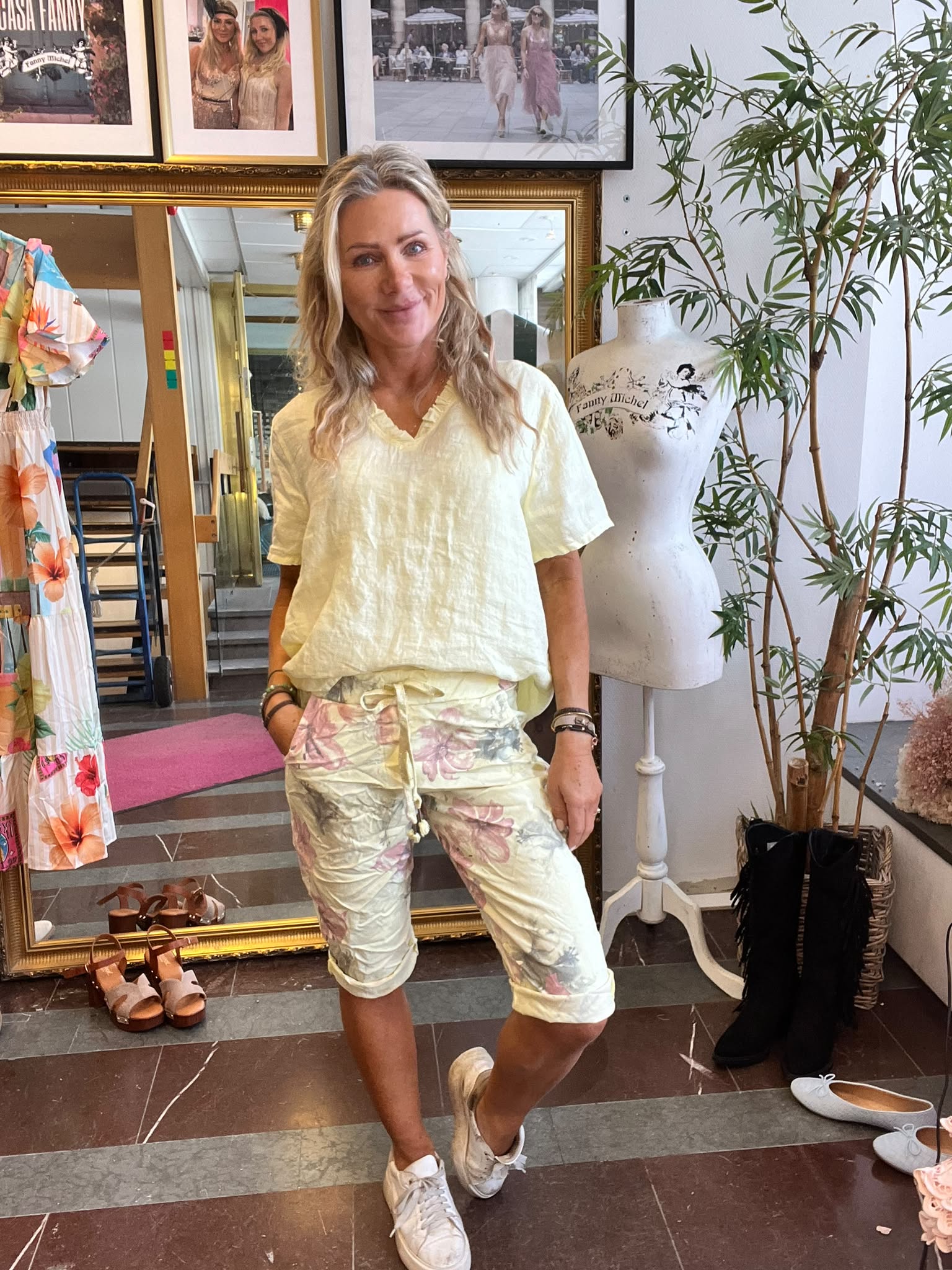 Blond kvinna står avslappnat och ler i en klädbutik, med händerna i fickorna, ljusgul topp och blommiga shorts. Bredvid syns provdocka med texten “Fanny Michel”, kläder, skor och stora speglar.