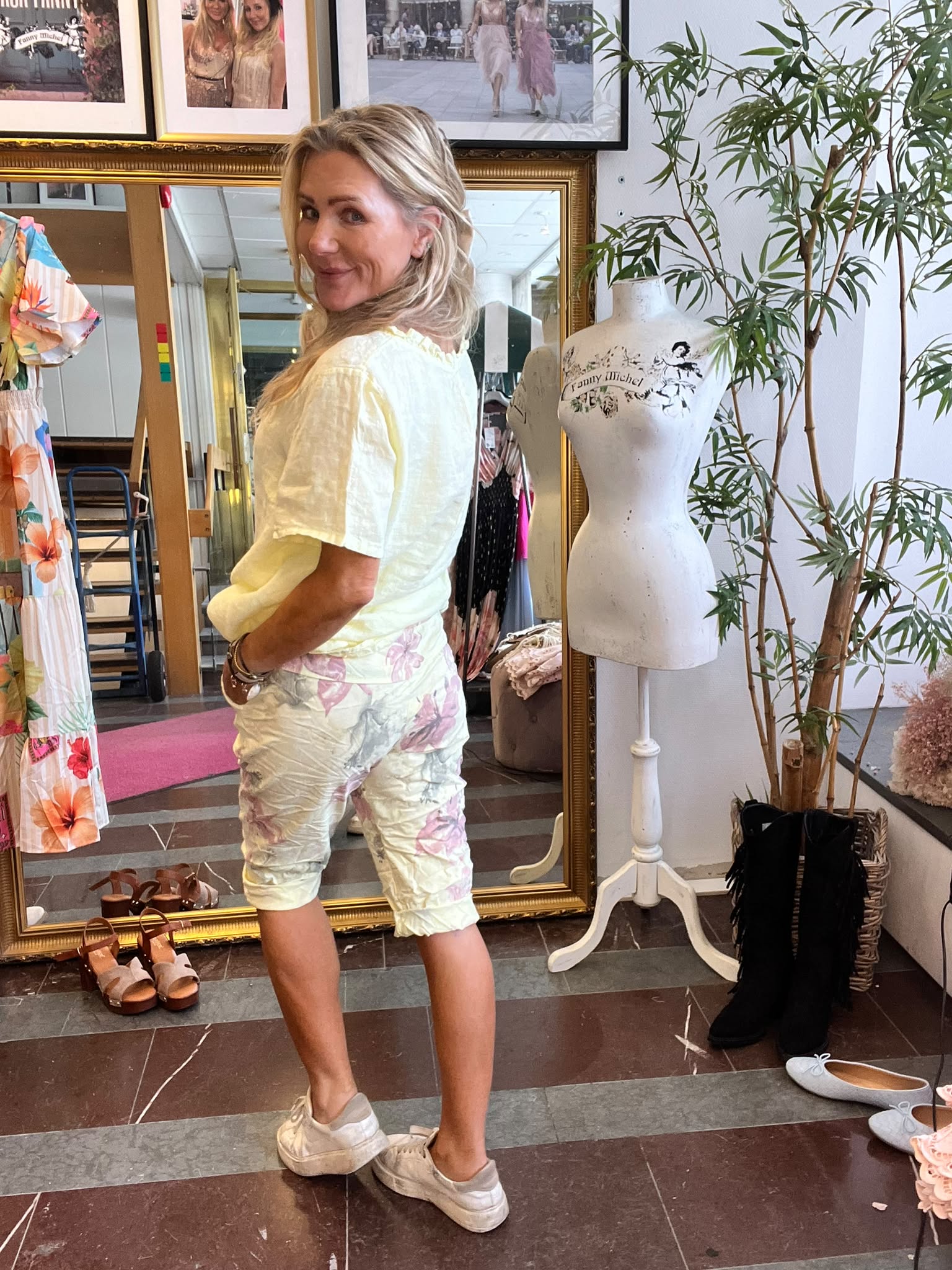 Leende kvinna vänder sig mot kameran med händerna i fickorna, klädd i ljusgul topp och blommiga shorts, i en klädbutik. Bredvid står en provdocka med texten “Funny Michel”, spegel och skor på golvet.