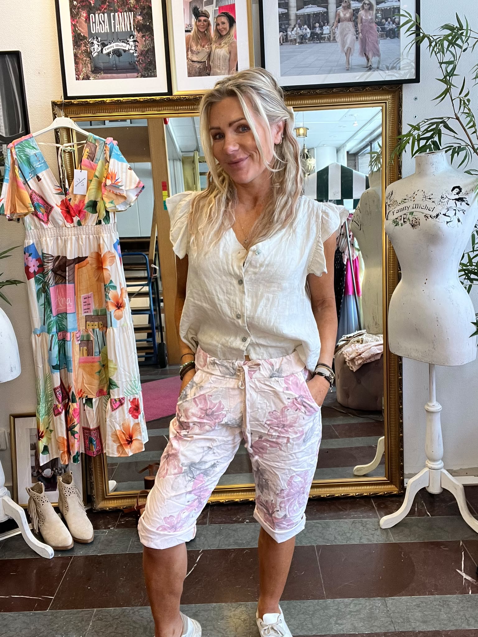 Blond kvinna står och ler med händerna i fickorna i en klädbutik, framför en stor guldramsspegel. Bredvid syns en färgglad klänning och en provdocka med texten “Fanny Michel”; på väggen “CASA FANNY”.