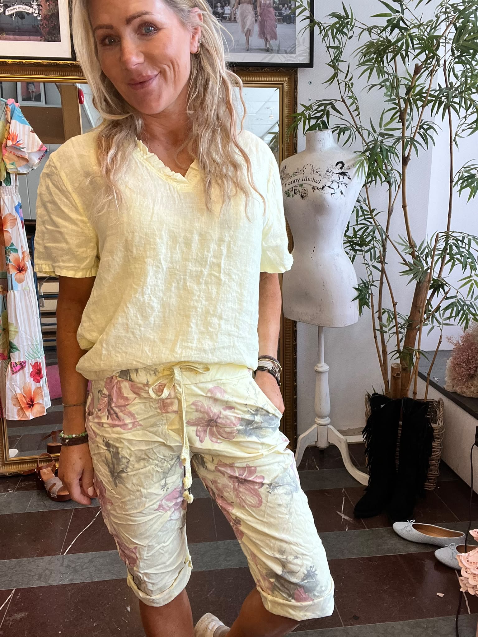 Leende kvinna poserar med handen i fickan och visar en ljusgul blus och blommiga capribyxor i en butik. I bakgrunden syns en provdocka med texten ”Fanny Michel”, speglar, kläder och växter.