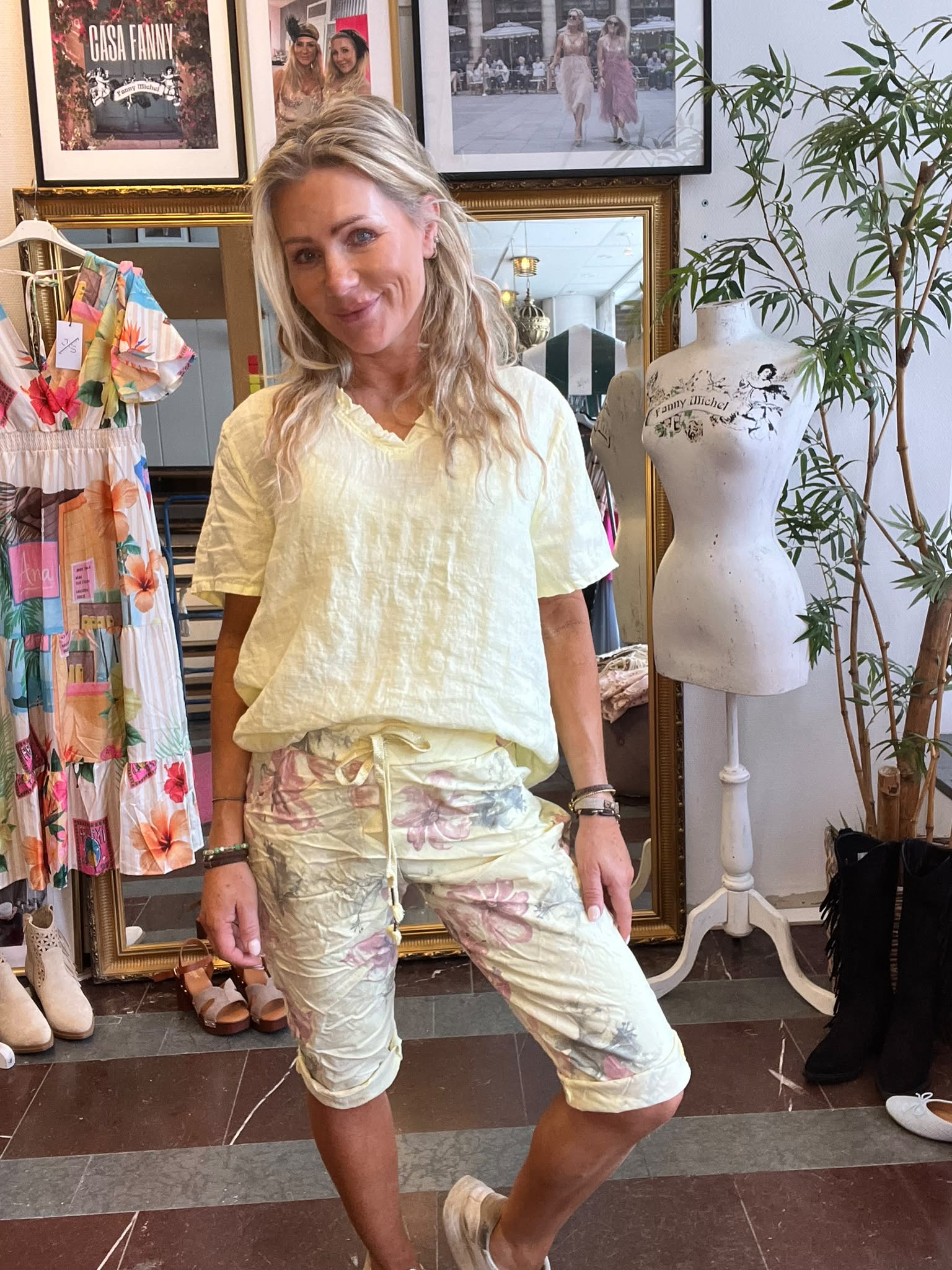Leende blond kvinna poserar i en klädbutik, iklädd ljusgul topp och blommiga capribyxor. Runt henne syns en färgstark klänning på galge, skor på golvet, en provdocka med texten “Fanny Michel” och en tavla med “CASA FANNY”.