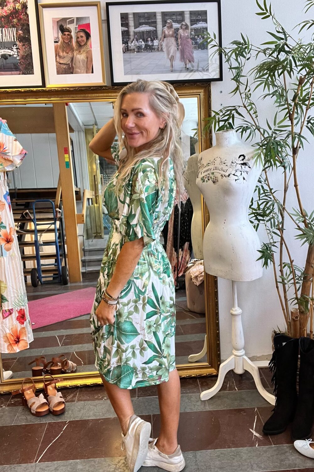 Kvinna i grönblommig klänning poserar och ler med handen i håret framför en stor guldramsspegel i en klädbutik. Runt henne syns klänningar, skor, provdocka med texten “Fanny Michel” och en skylt “CASA FANNY”.