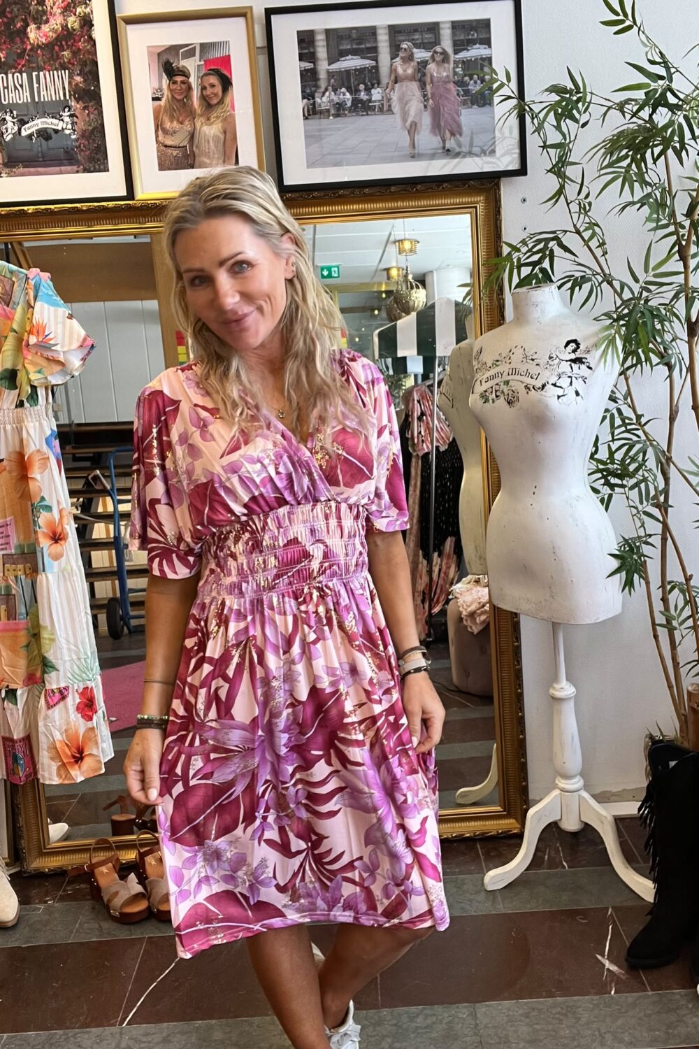 Leende kvinna i rosa mönstrad klänning står i en klädbutik och håller en solglasögon i handen. Runt henne syns klänningar, skor, en provdocka och inramade foton med texten “CASA FANNY” och “Fanny Michel”.