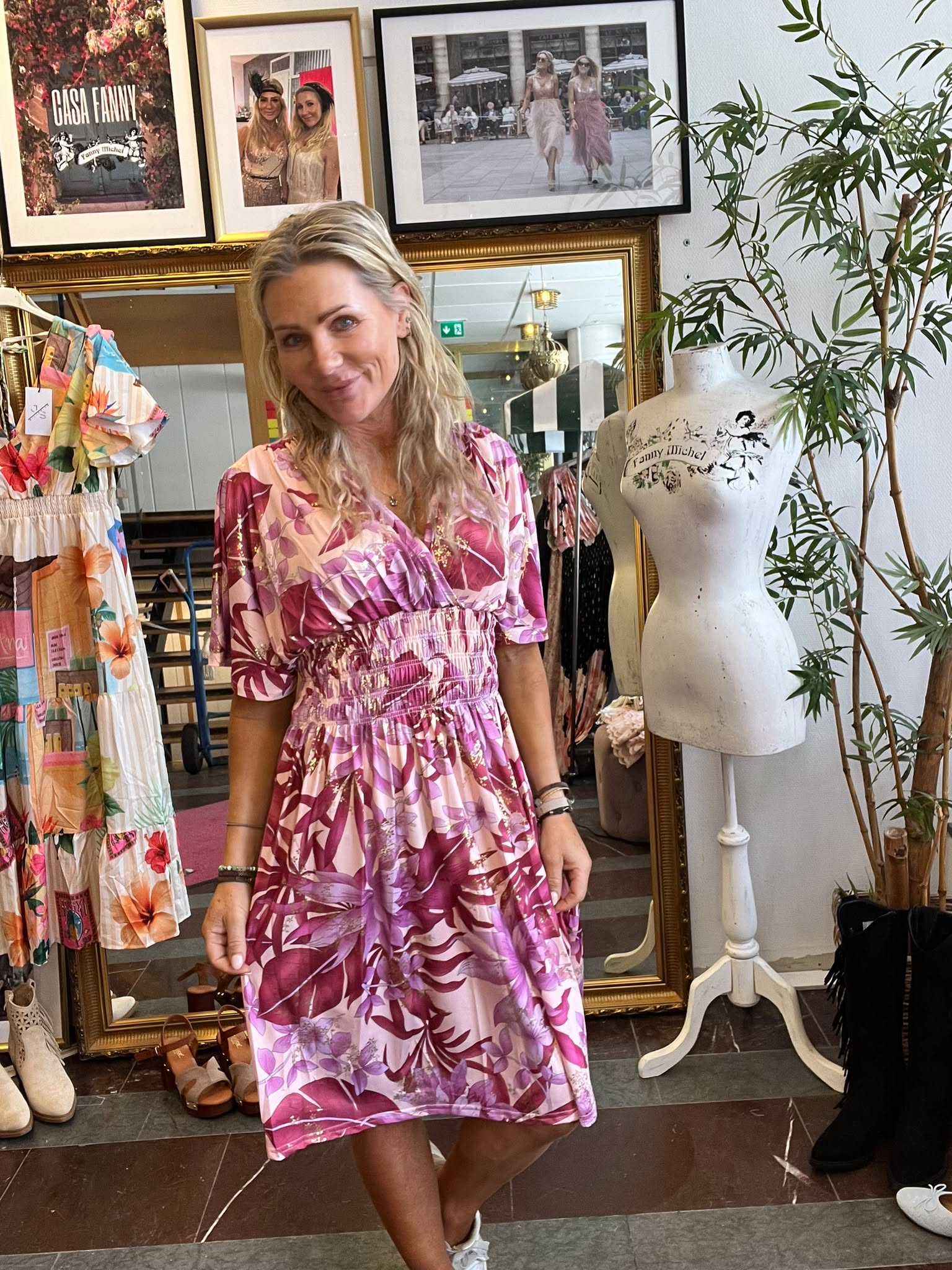 Leende kvinna i rosa mönstrad klänning står i en klädbutik och håller en solglasögon i handen. Runt henne syns klänningar, skor, en provdocka och inramade foton med texten “CASA FANNY” och “Fanny Michel”.