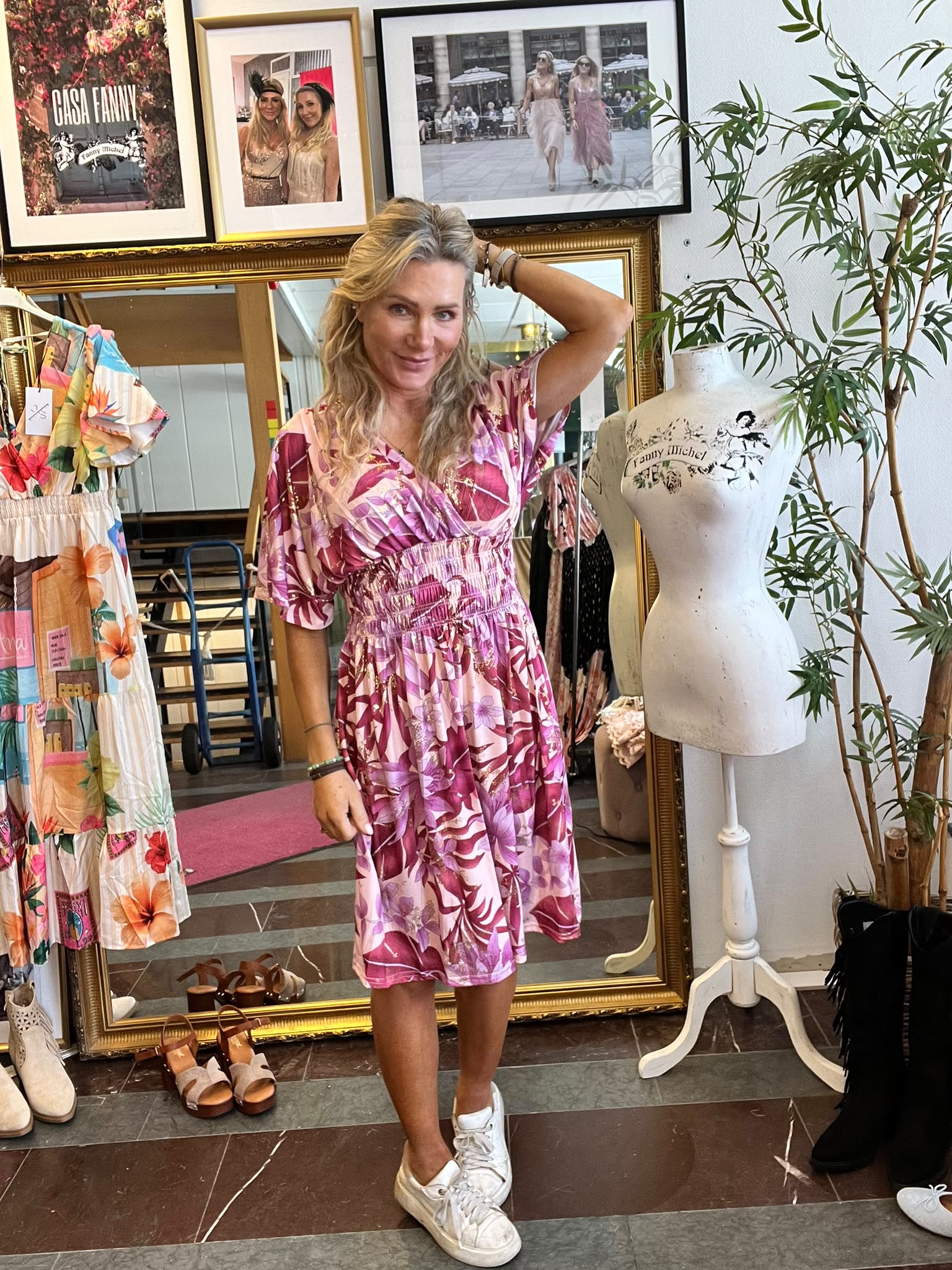 Kvinna poserar och ler med ena handen i håret, klädd i rosa blommig klänning och vita sneakers, i en klädbutik. Bredvid står provdocka med texten “Fanny Michel”; bakom syns spegel, klänningar och tavlor, inklusive “CASA FANNY”.