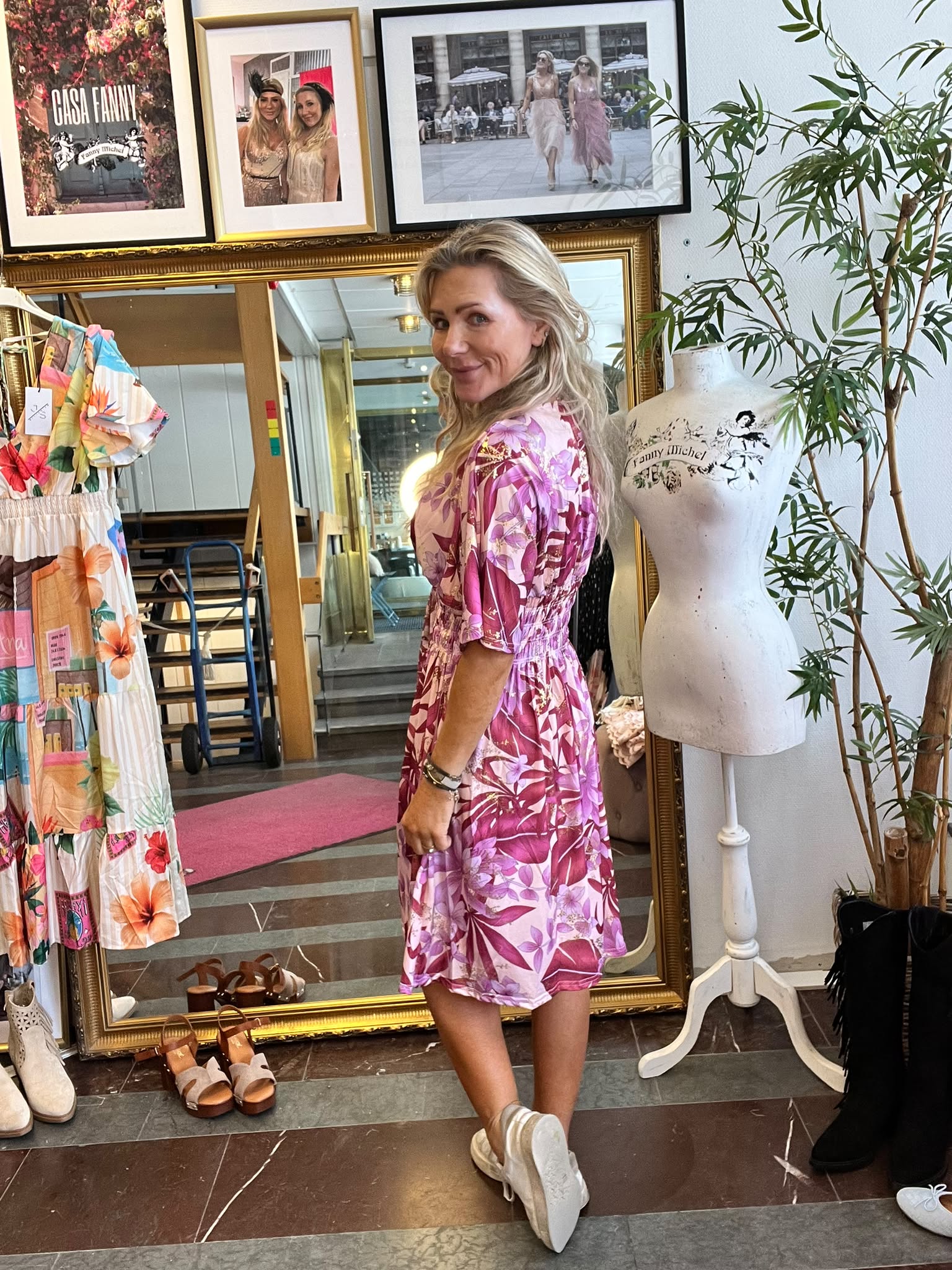 Kvinna i rosa, blommig klänning vänder sig om och ler mot kameran i en klädbutik. Bredvid står en provdocka med texten ”Fanny Michel”; bakom syns guldramsspegel, klänningar och inramade foton.