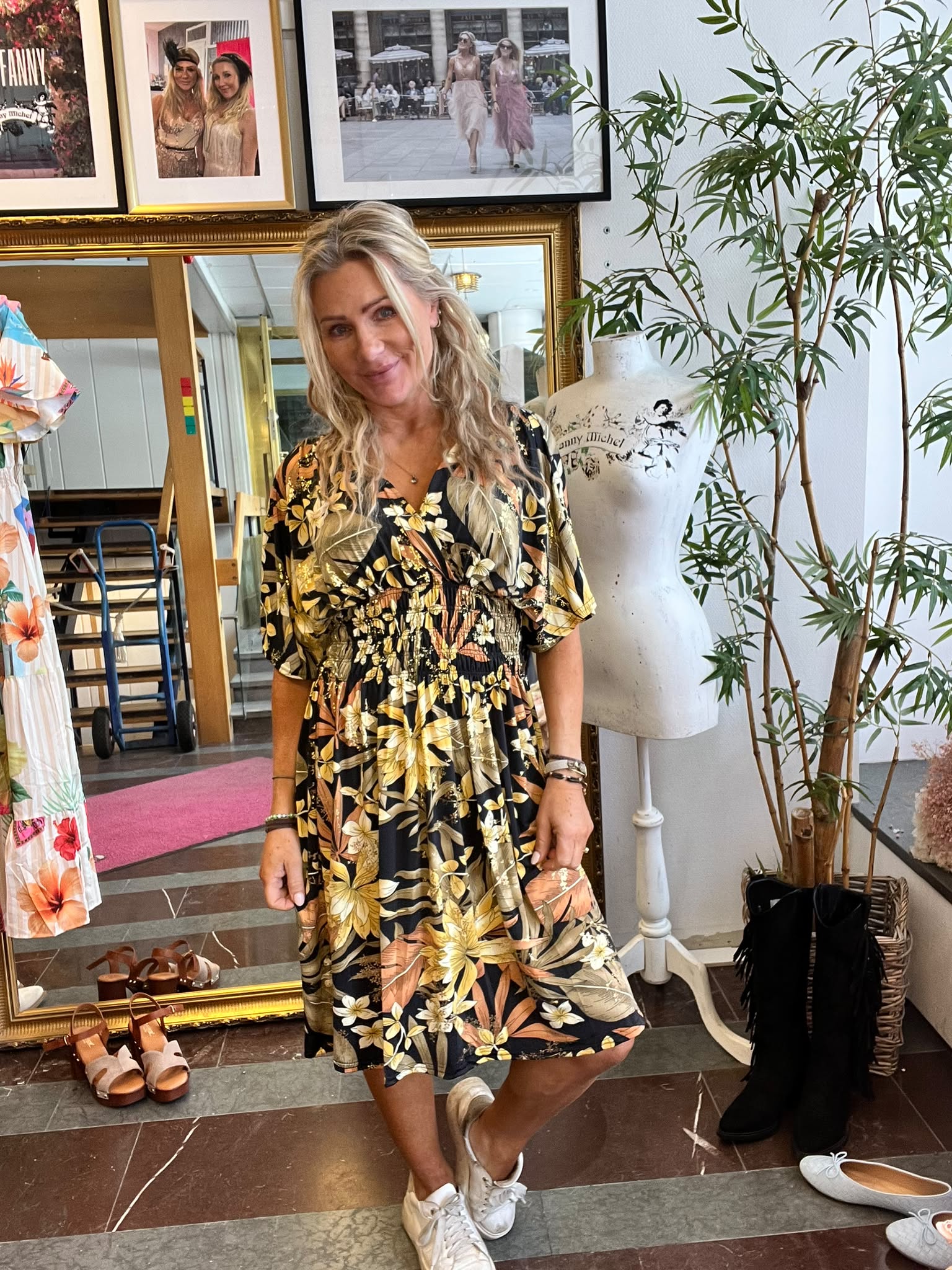 Leende kvinna poserar i en butik/ateljé, klädd i svart blommig klänning och sneakers. Hon står vid en provdocka med texten “Ginny Michel”, omgiven av skor på golvet, speglar, inramade foton och en hög växt.