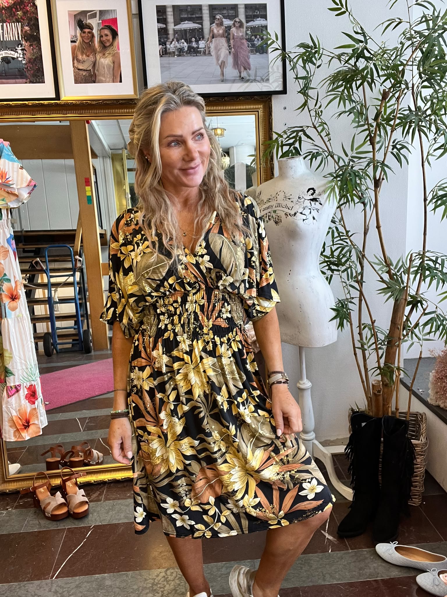 Kvinna i svart, blommig klänning poserar leende och lyfter lätt i kjolen i en butik. Runt henne syns skor på golvet, en provdocka med texten “Fanny Michel”, inramade foton och en stor grön växt.