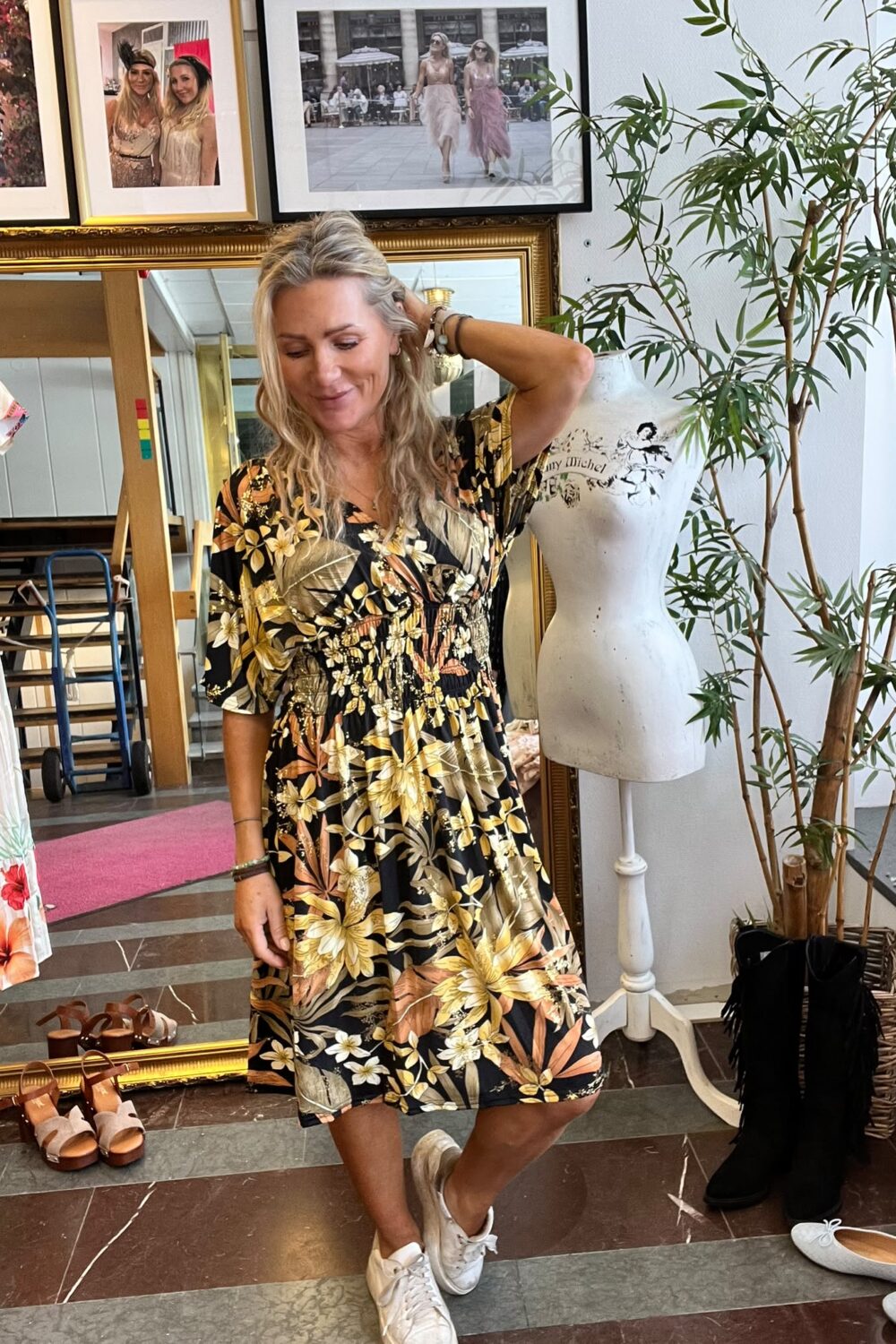 Blond kvinna provar en svart blommig klänning och står avslappnat med handen i håret. Hon är i en butik/ateljé med kläder och skor, spegel, provdocka med texten ”Fanny Michel” och inramade foton på väggen.