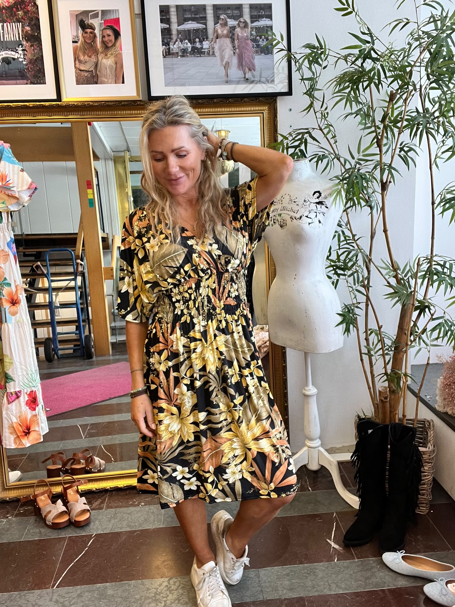 Blond kvinna provar en svart blommig klänning och står avslappnat med handen i håret. Hon är i en butik/ateljé med kläder och skor, spegel, provdocka med texten ”Fanny Michel” och inramade foton på väggen.