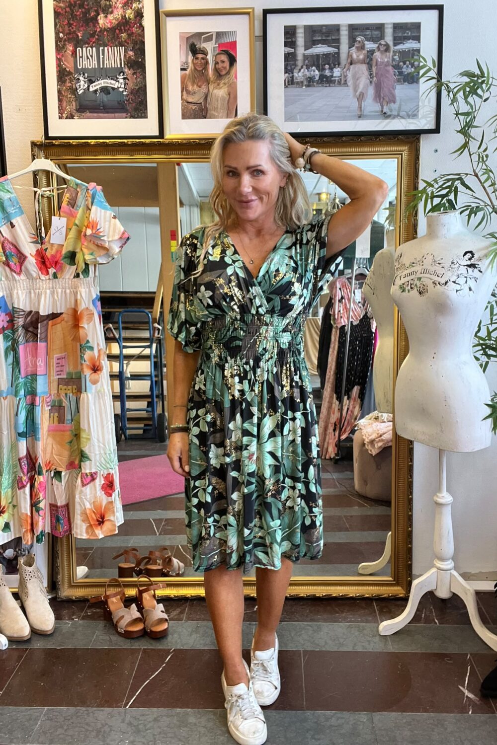Leende kvinna poserar med ena handen i håret i en klädbutik, iklädd grön blommig klänning och vita sneakers. Runt henne syns klänningar, skor och provdocka; ovanför hänger inramade foton med texten “CASA FANNY”.