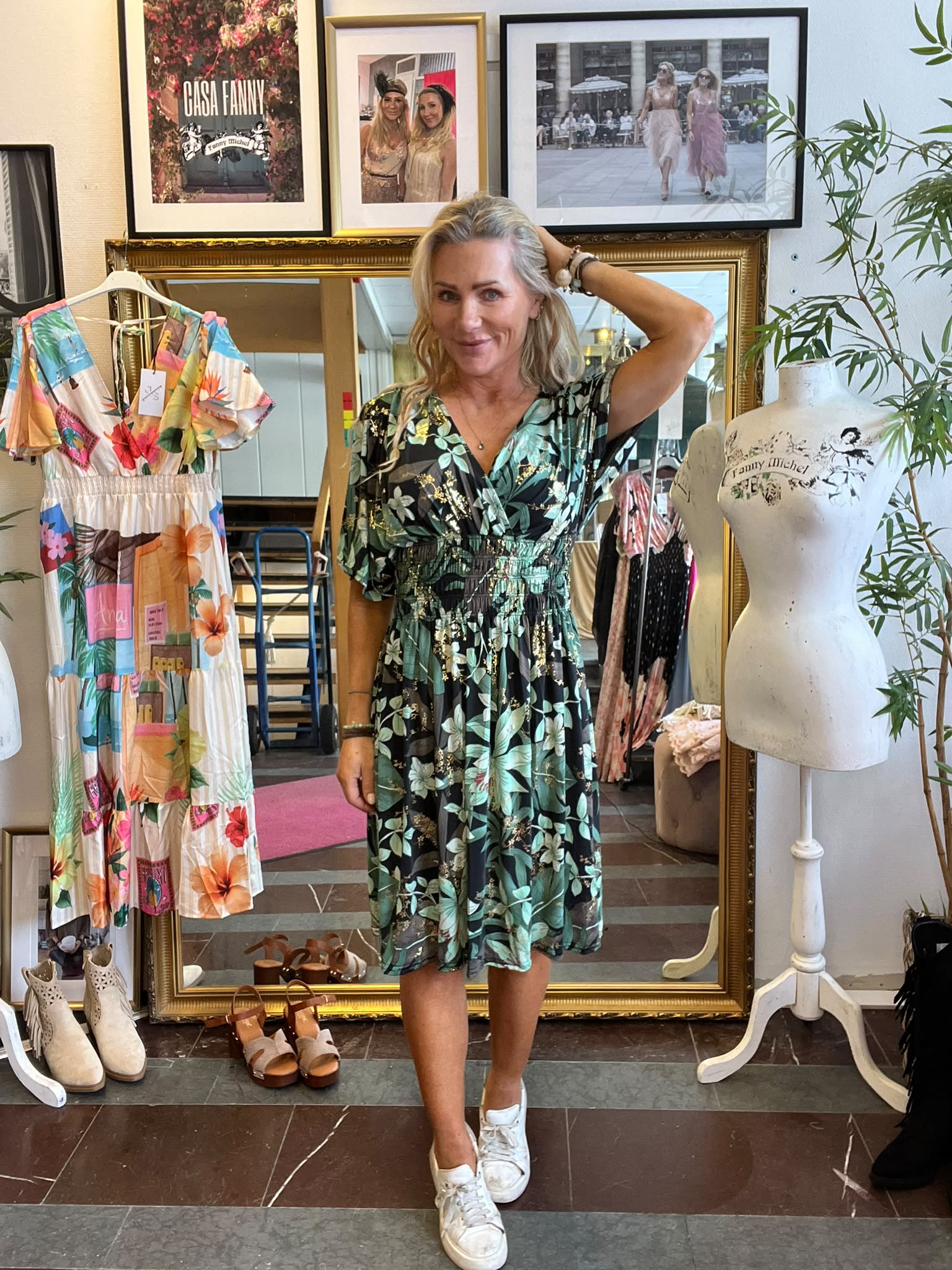 Leende kvinna poserar med ena handen i håret i en klädbutik, iklädd grön blommig klänning och vita sneakers. Runt henne syns klänningar, skor och provdocka; ovanför hänger inramade foton med texten “CASA FANNY”.