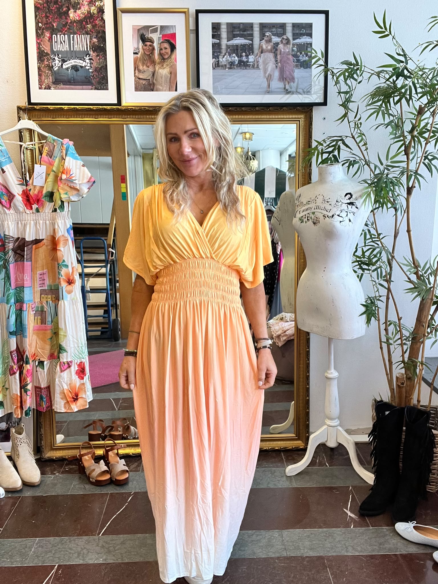 Kvinna står och poserar i en ljusorange långklänning i en klädbutik. Bakom henne syns en stor guldramsspegel, en skyltdocka med texten ”Fanny Michel”, skor på golvet och inramade foton, bland annat ”CASA FANNY”.