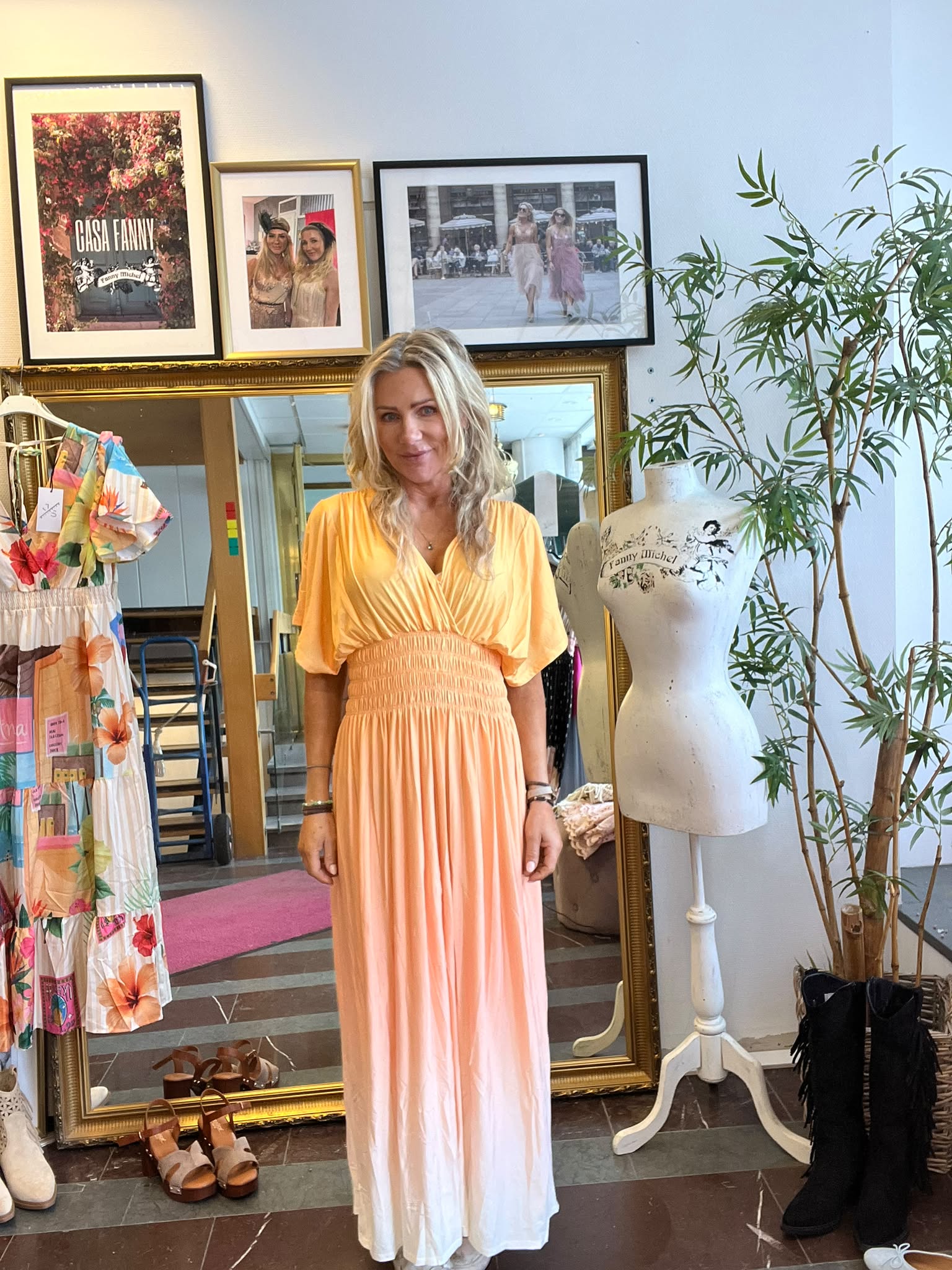 Kvinna i gul‑orange långklänning står och ler i en klädbutik framför en stor spegel. Runt henne syns klänningar, skor och en provdocka med texten “Fanny Michel”, samt inramade foton inklusive “CASA FANNY”.