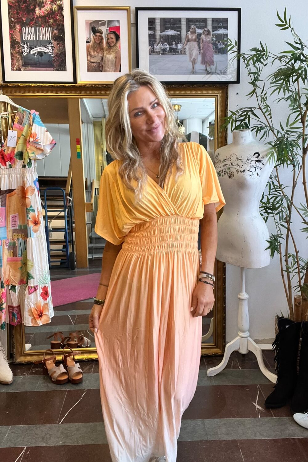 Leende kvinna i en lång persikofärgad klänning står och poserar i en butik. Runt henne syns en blommig klänning på galge, skor på golvet, provdocka med texten ”Fanny Michel” och inramade foton, inklusive ”CASA FANNY”.